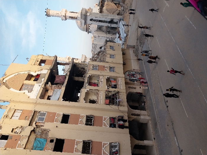 The image captures a street scene in Khan Yunis, Palestinian Territory, displaying widespread damage to urban structures. A multi-story residential building, light beige in color with decorative arched window frames, dominates the left side, exhibiting extensive structural damage. Large sections of its exterior walls are missing, revealing interior spaces and debris. Numerous windows are broken or entirely absent. Clothes hang from railings on several damaged balconies. In the background, a light-colored mosque with a prominent minaret and dome is visible, also appearing heavily damaged with collapsed sections and surrounding rubble.
The paved street in the lower portion of the image contains white lane markings and scattered debris. Approximately 20-30 individuals, primarily children and young adults, are present. Several groups are seen walking across the street, while others stand or gather on the sidewalk near the damaged residential building's arched ground-floor openings. The scene is illuminated by daylight under a partly cloudy blue sky.