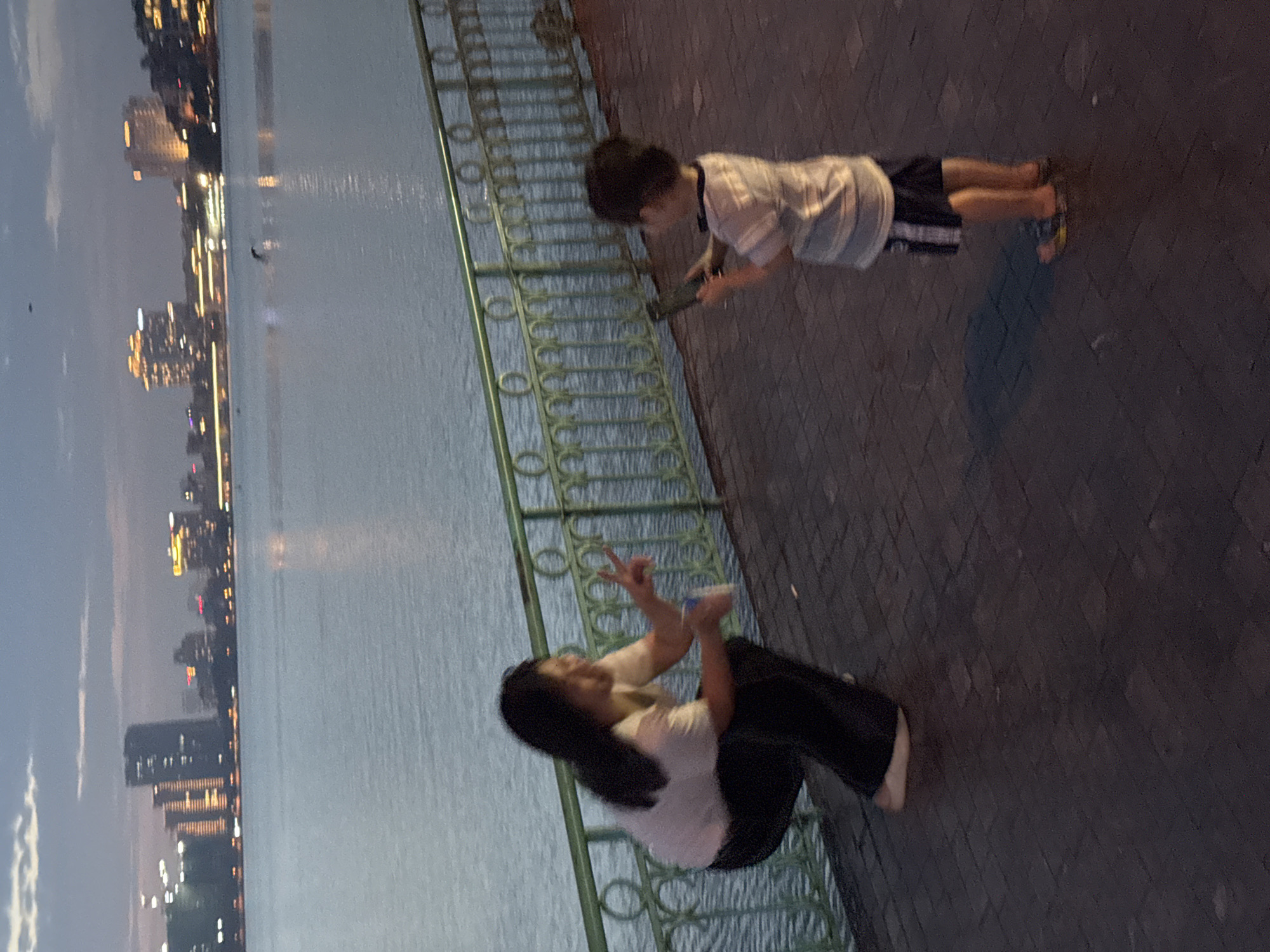 The image depicts a woman and a young boy on a paved walkway beside a body of water, likely a lake or river, with a city skyline in the background. The woman is crouched down, wearing a white t-shirt and black pants. She is holding a white cup and making a peace sign with her right hand. The boy, facing away from the camera, is wearing a striped t-shirt and dark shorts. He is holding a dark object, possibly a phone, in his hands. A green metal railing separates the walkway from the water. The city skyline features numerous buildings, some illuminated with lights, suggesting it is dusk or evening. The environment appears to be an urban park or waterfront area. The context of Hà Nội, Vietnam, suggests this scene could be along Hồ Tây (West Lake) or another prominent waterway within the city, given the characteristic urban skyline with illuminated buildings.