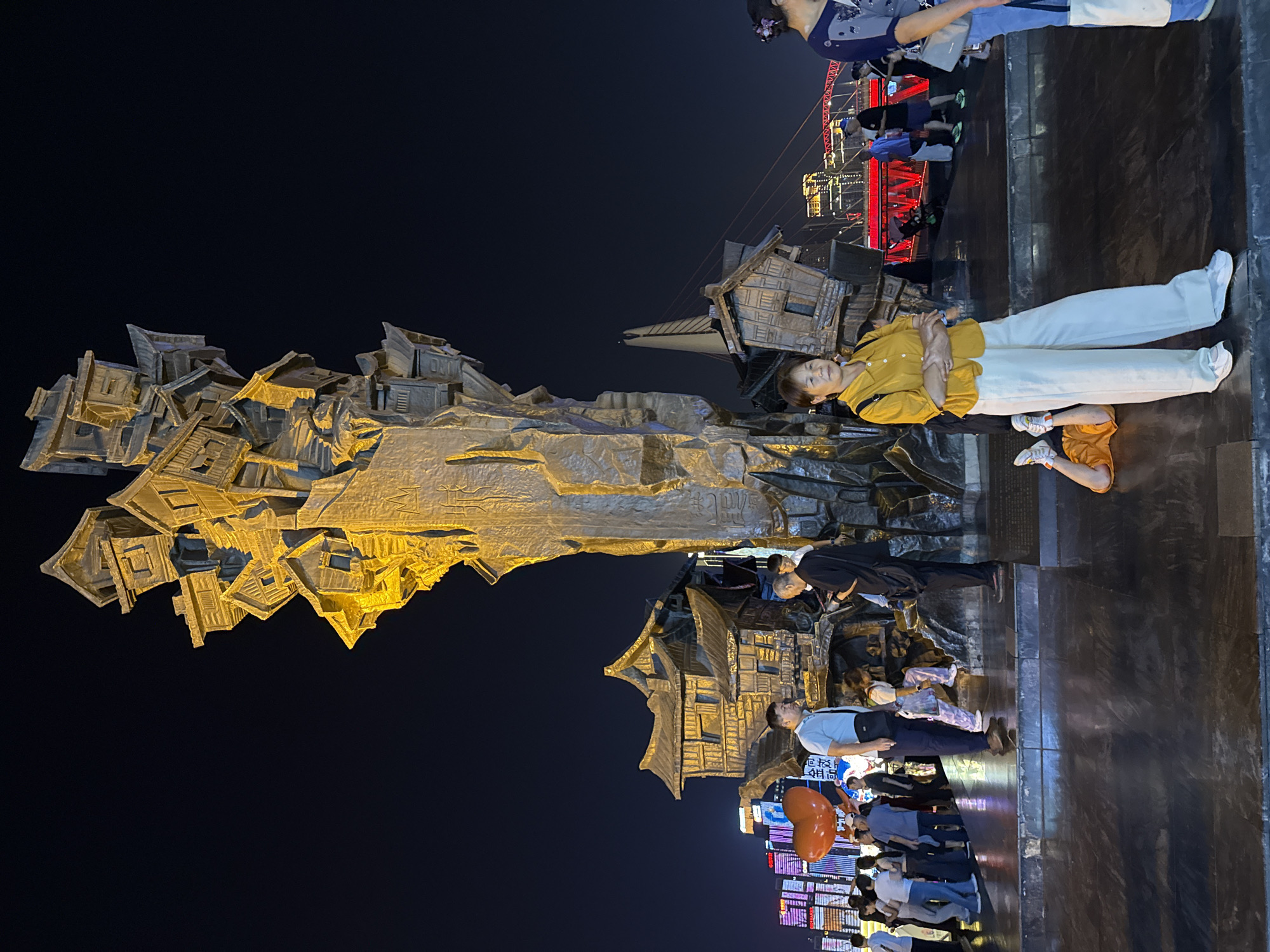 The image depicts an outdoor scene in the Yuzhong District of Chongqing, China, at night. A large, abstract sculpture resembling a collection of traditional Chinese houses dominates the left side, illuminated with golden light. People are gathered around the sculpture, with some interacting with it. A woman in a yellow shirt and white pants poses directly in front of the sculpture. A pedestrian bridge with red railings and decorative lights is visible in the background. The ground is paved with dark, reflective tiles, mirroring the surrounding lights and structures. Illuminated buildings are present along the bottom of the image, suggesting a lively urban environment.