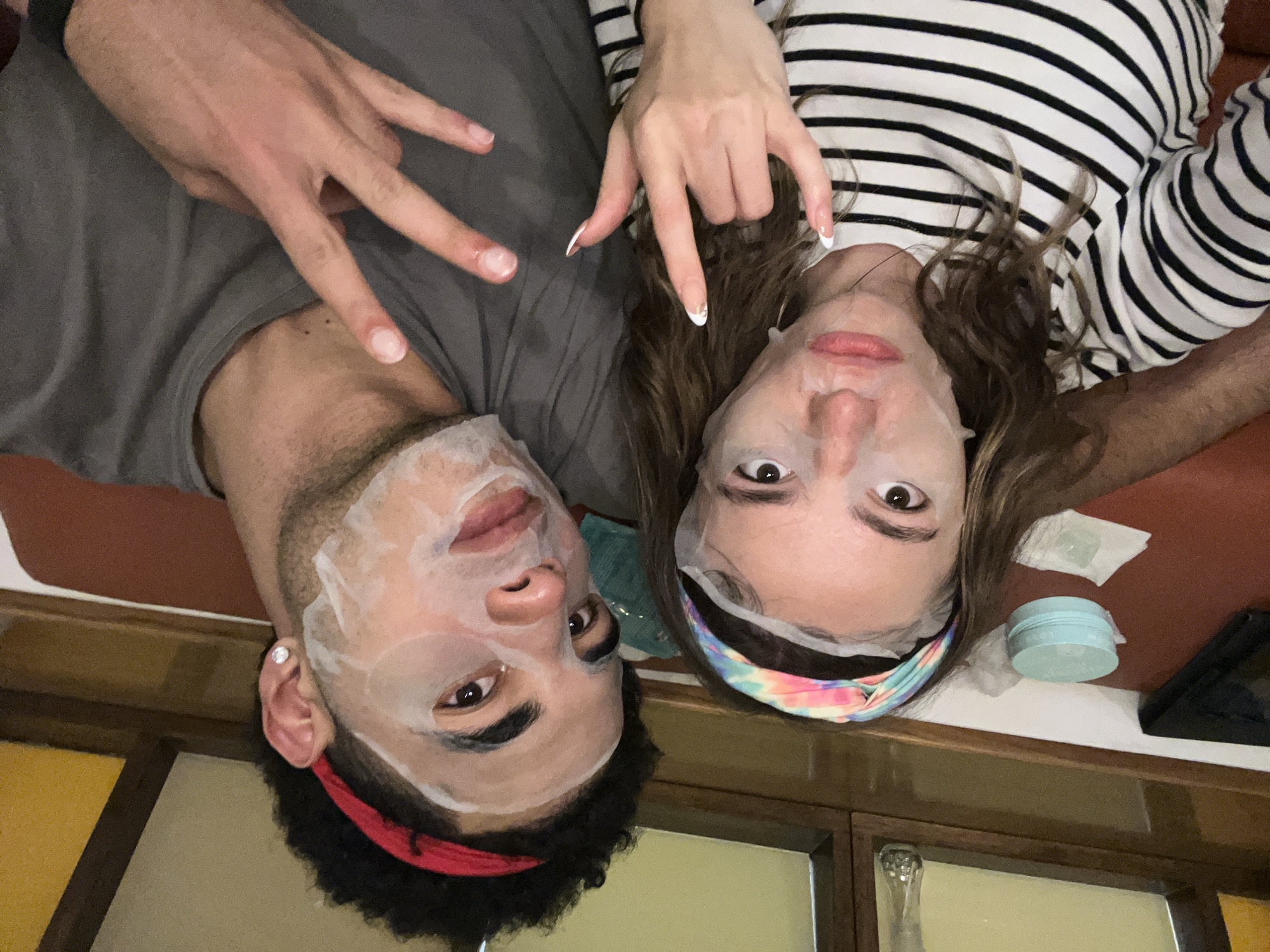 A medium shot depicts two individuals, a male and a female, lying on their backs with facial masks applied. The male is positioned on the left, wearing a gray t-shirt and a red headband, and he is making a hand gesture with his left hand. The female, on the right, is wearing a black and white striped top and a colorful headband, also making a hand gesture with her right hand. A small container of skincare product and tissues are visible near the female's head. The location appears to be indoors, possibly a bathroom, with a wood-paneled cabinet visible in the background. Specific details suggesting the city of San Bernardino Tlaxcalancingo, Mexico are not discernible from the image.