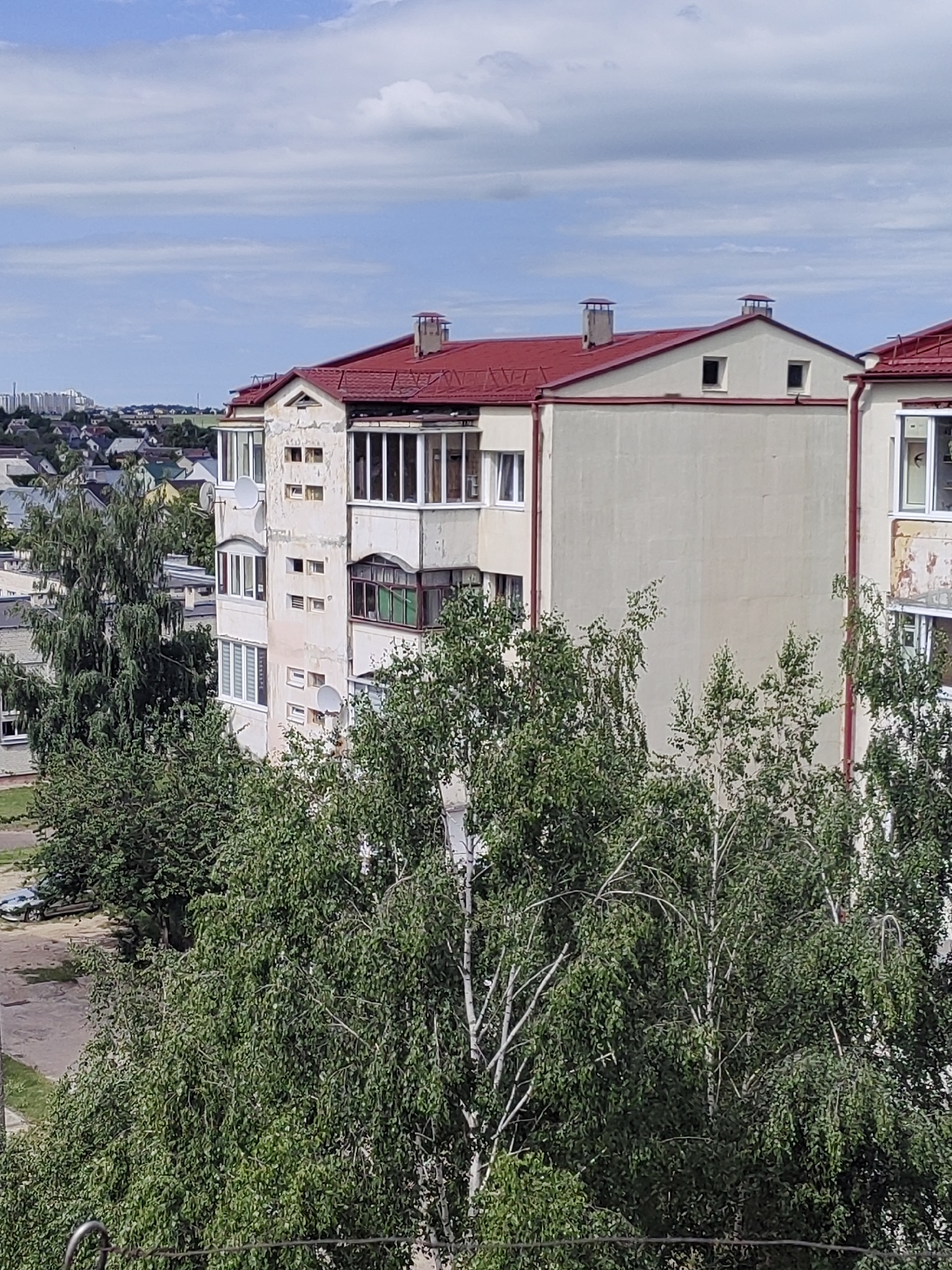 This is an outdoor, eye-level shot of a multi-story apartment building with a red roof, taken from above. The building appears to be made of concrete and has a light-colored facade. It is partially obscured by lush green trees in the foreground. The building has several windows, some of which are balconies with glass enclosures. There are satellite dishes mounted on the side of the building. In the background, other buildings and trees are visible, with a cityscape in the distance. The sky is blue with scattered white clouds, suggesting it is daytime and the weather is fair. The scene appears to be a residential area. There is no text visible in the image.