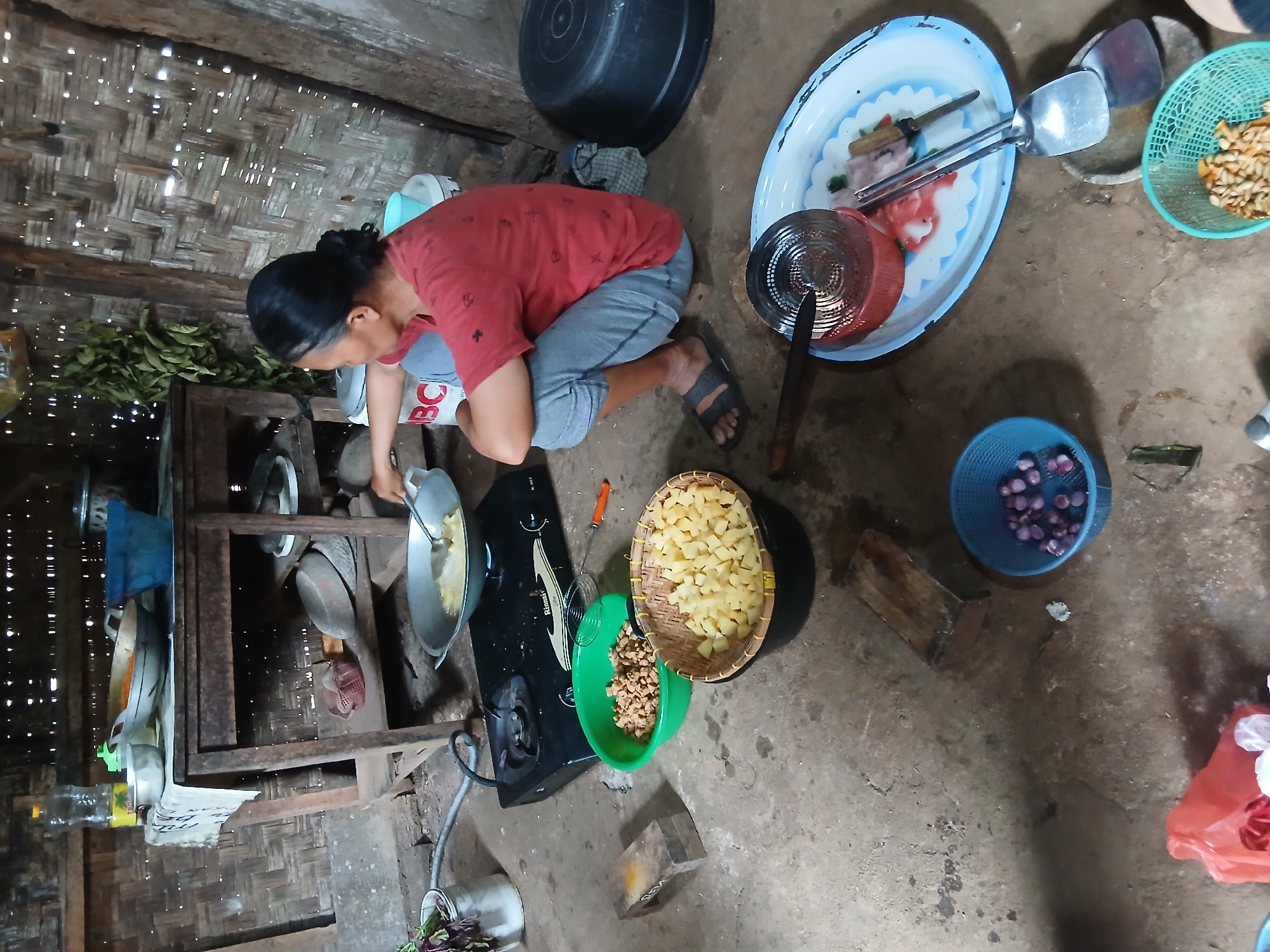 A woman wearing a red t-shirt and gray pants is crouching on the floor, cooking in a wok on a gas stove. The scene takes place indoors, in what appears to be a simple kitchen. The walls are made of woven bamboo, and the floor is concrete. There are several bowls and containers scattered around, filled with ingredients such as chopped potatoes and small pieces of what look like fried snacks or pork rinds. A large black plastic basin is visible to the right, and a blue plastic basket containing onions is in the foreground. A table made of wood stands behind the stove, with various cooking utensils and bowls on it. A bottle of cooking oil is also on the table. The overall impression is that of a domestic scene in a rural or humble setting in Kalirejo, Indonesia.