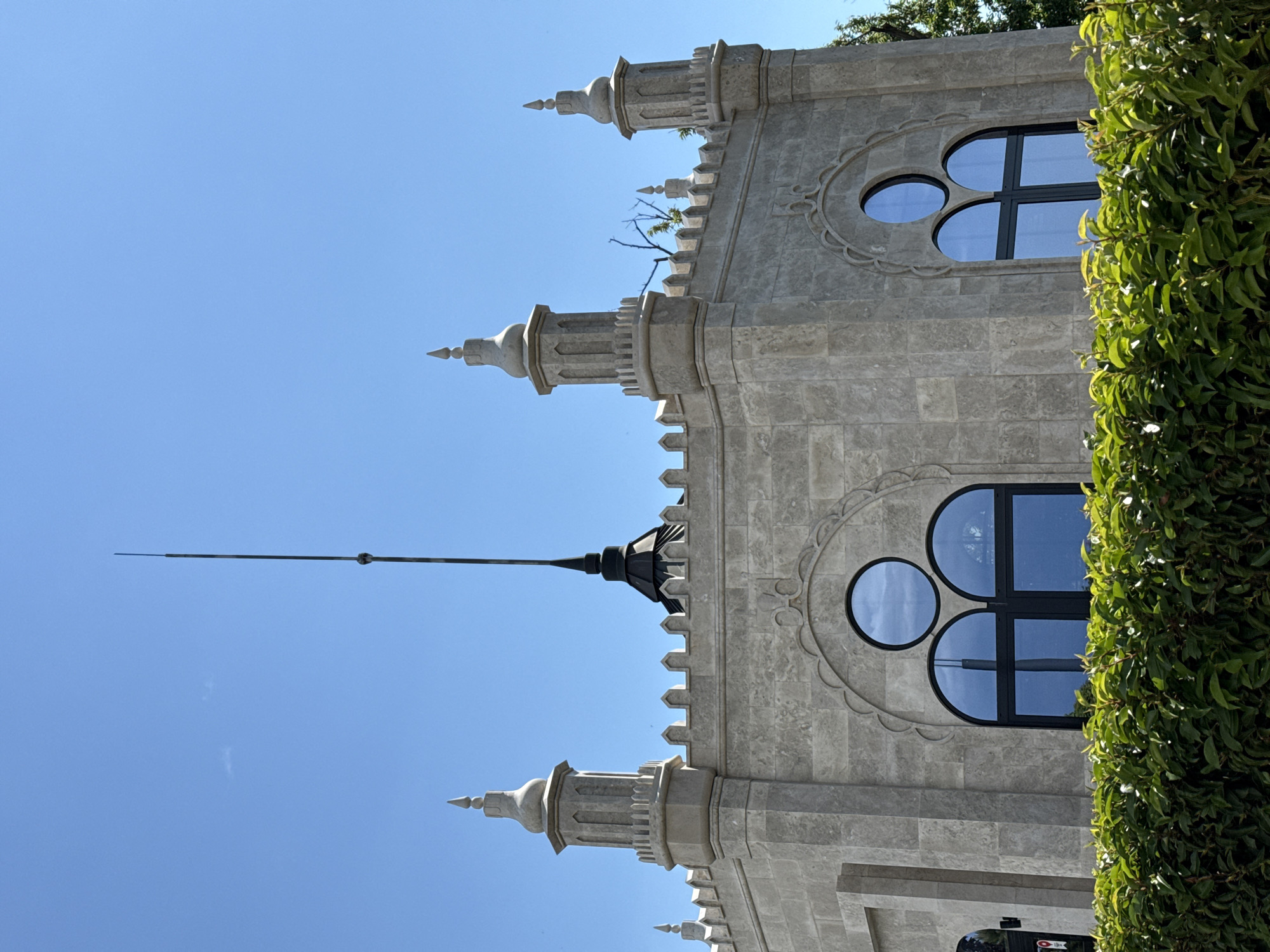 The image shows the upper portion of a light-colored stone building with an octagonal shape, featuring decorative crenellations along the roofline and four ornate towers. A prominent black lightning rod extends vertically from the central roof peak. Two large, dark-framed windows, each composed of two arched panes topped by a circular pane, are visible. A dense green hedge runs along the bottom of the frame. The sky is a clear, bright blue. The scene is set outdoors, in a location that is not identifiable from the image.
