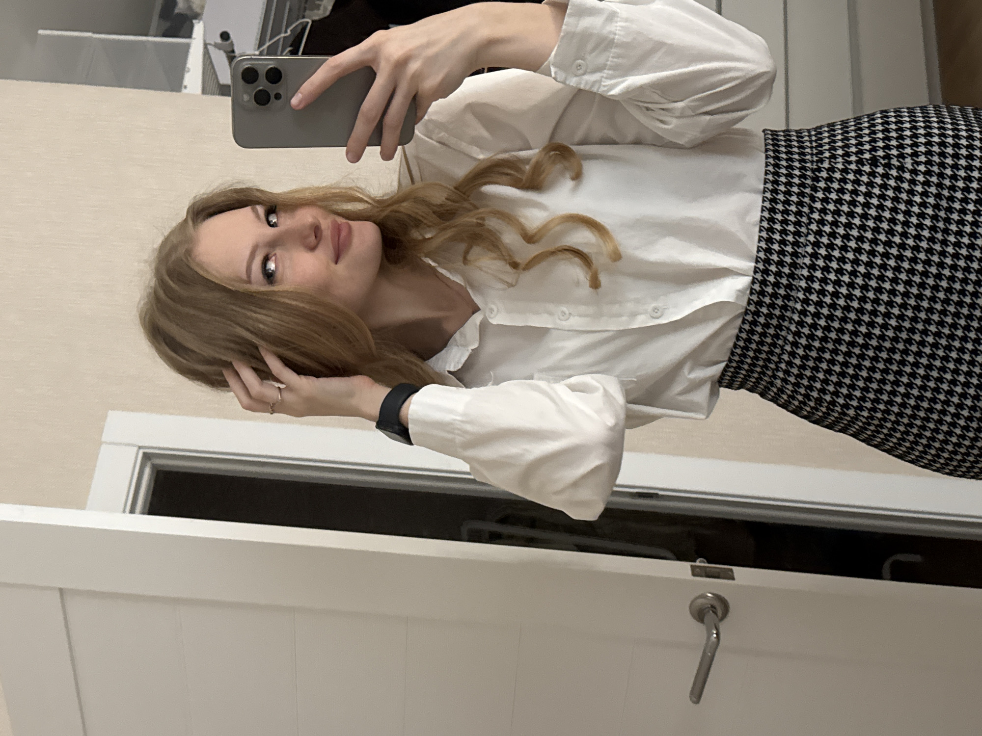 A young woman with blonde hair, wearing a white button-down shirt and a black and white houndstooth skirt, takes a selfie in a room. She is holding a gray smartphone in her right hand and touching her hair with her left. A watch is visible on her left wrist. The background includes a white door with a silver handle on the left, a doorway with a dark interior, and a wall with a textured pattern. There are also some shelves visible in the reflection of the phone. The location is not specified in the image, and therefore, no location in the city of Perm, Russia, can be determined.