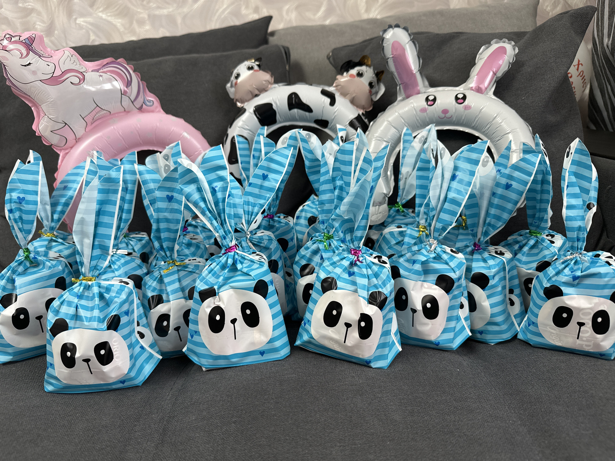 The image shows a collection of panda-themed party favor bags. The bags are light blue with horizontal stripes and a panda face design on each. They are gathered at the top and tied with metallic string. In the background, there are three mylar balloons: a pink unicorn balloon, a cow balloon, and a white rabbit balloon. The bags and balloons are arranged on a gray, textured surface, possibly a sofa or other soft furnishing. The location is Chaikovsky, Russia.