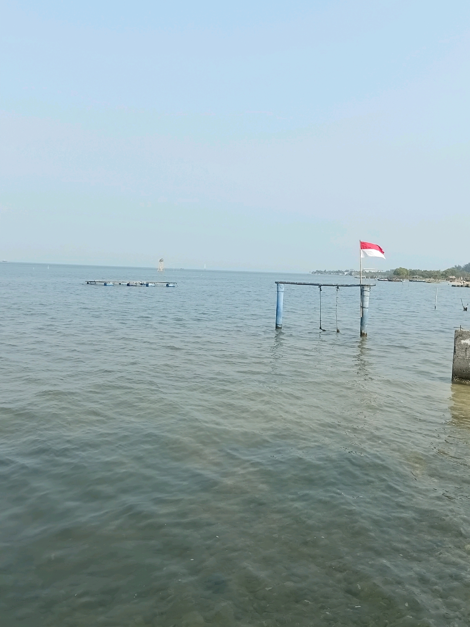 The image depicts a tranquil body of water, likely a sea or large lake, under a clear, pale blue sky. In the foreground, the water is a muted green, with subtle ripples indicating a gentle breeze. Partially submerged in the water are two blue-painted posts connected by a horizontal bar, with two ropes hanging down, suggesting a swing set structure. A red and white flag, the national flag of Indonesia, is flying from the top of the structure.
Further in the distance, a floating platform or raft made of buoyant barrels and a rectangular frame is visible. Beyond that, a lighthouse-like structure stands on a small island or platform in the water. The shoreline, lined with vegetation and some man-made structures, can be seen in the far distance on the right side of the frame. The overall scene suggests a coastal or lakeside area in Cikal, Indonesia, characterized by calm waters and open skies. There are no people or animals visible in the image. The lighting suggests it is daytime, likely mid-morning or afternoon.