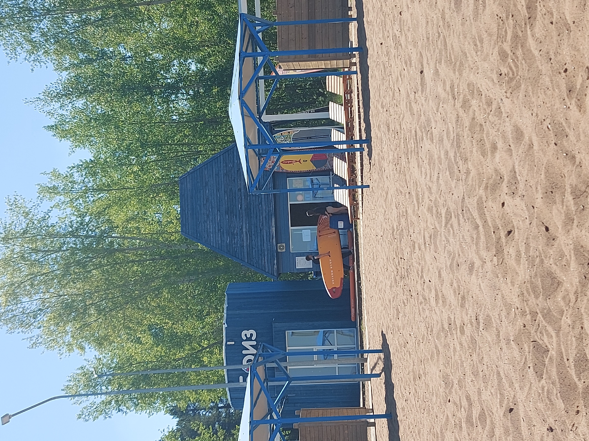 The image depicts a beach in Chaikovsky, Russia. The scene includes a sandy beach, blue buildings with white windows, and blue metal shade structures. A paddleboard rests against one of the buildings. Green trees line the background.