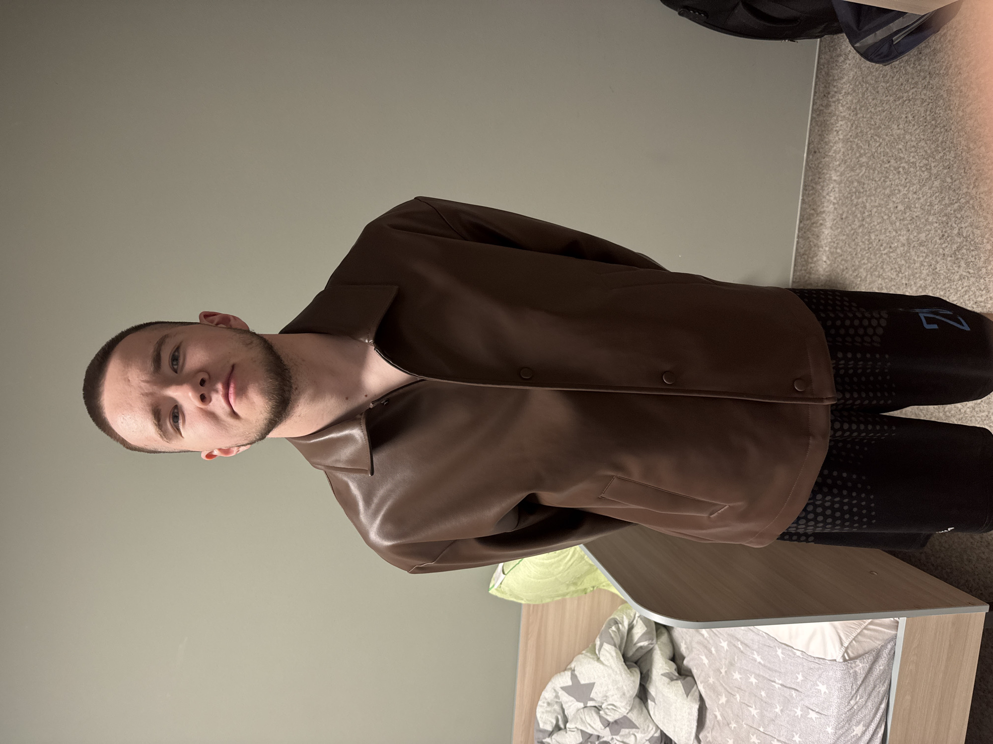 A young man is featured in this indoor portrait, likely taken in a bedroom or dorm room in Kazan, Russia. He is positioned facing the camera, with a direct and neutral gaze, and has short dark hair and a trimmed beard.
He is wearing a brown, collared jacket made of a material resembling leather or faux-leather, secured with dark buttons down the front. Below the jacket, he wears dark shorts or athletic pants adorned with a pattern of light grey dots and a distinctive blue graphic on the side, which appears to be a stylized "VZ" or "7".
The background shows a simple room setup. A plain light grey wall is on the left, while the floor on the right is covered with a speckled grey carpet. A wooden bedside table or headboard is visible in the lower right corner, next to a bed dressed with a grey and white star-patterned duvet and a light green pillow. A dark backpack or bag can be seen partially in the upper right background. The room appears to be lit by artificial light. The image is presented with a 90-degree clockwise rotation.