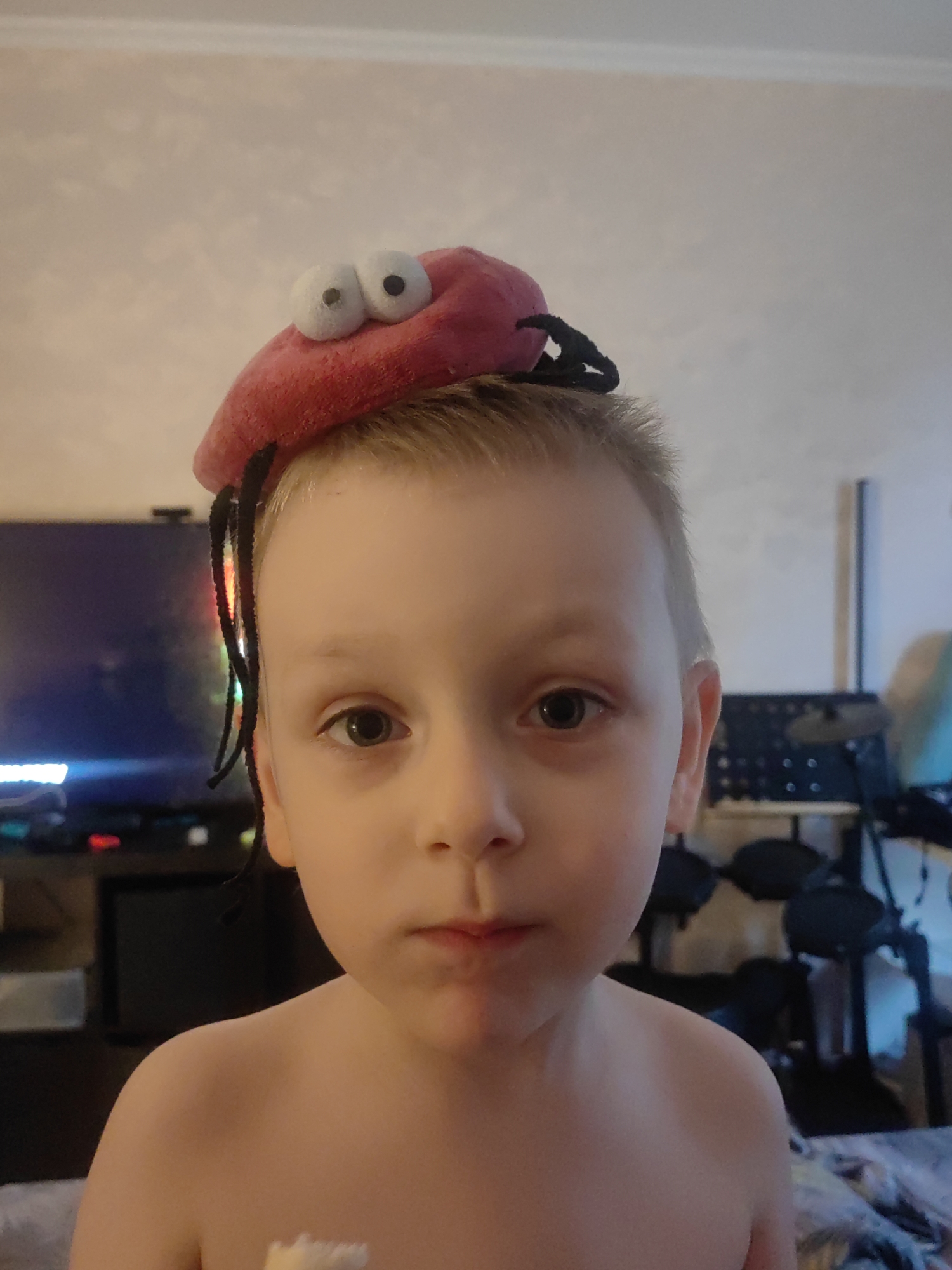 A close-up shot depicts a young, light-skinned boy with blond hair and light-colored eyes. He has a serious expression and is wearing a novelty hat resembling a red bug with large, googly eyes and black antennae. His shoulders are bare, suggesting he may be shirtless. The background includes a television and what appears to be a musical keyboard and drum set. The exact location in the city of Mytishchi, Russia, cannot be determined from the image.