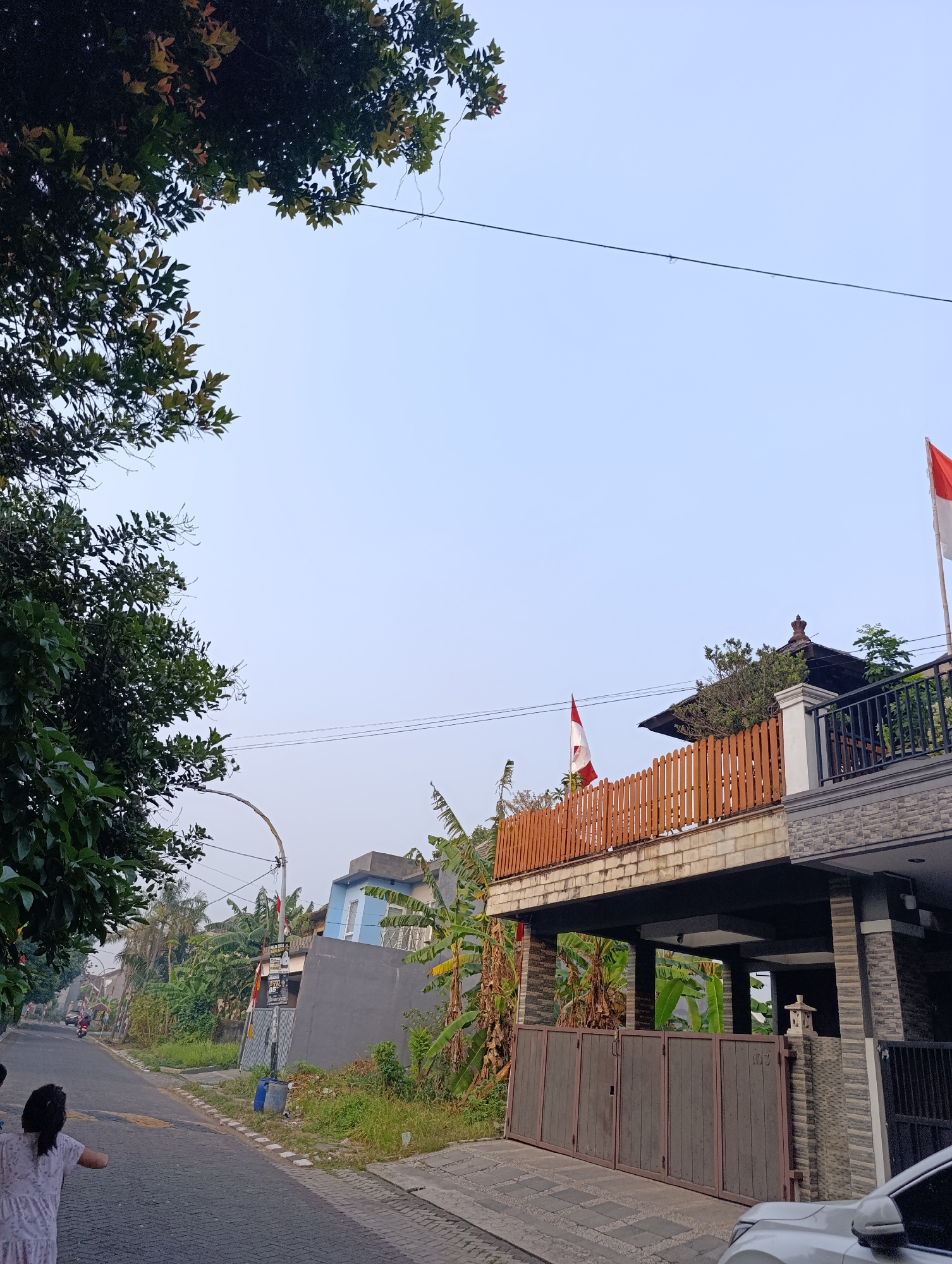 The image shows a residential street in Tangerang, Indonesia, under a clear, pale blue sky. On the left, a large tree with dark green leaves partially obscures the sky. The street, paved with dark interlocking bricks, curves gently to the left and is lined with houses and lush vegetation, including banana trees. A street lamp with a bent pole is visible along the roadside.
In the foreground on the left, a young person wearing a light-colored dress stands with their back to the camera, facing away from the viewer and towards the street. To the right, a multi-story house is prominent, featuring a balcony with a light brown wooden railing. Two Indonesian flags are displayed on the house: one on the balcony and another higher up on the right side of the building. The house has a stone-tiled facade and a dark-colored gate. A white car is parked in front of the house on the right side of the image. In the middle ground, several other houses and buildings, some with blue walls, are visible amidst dense greenery. A motorcycle is seen on the street in the distance.