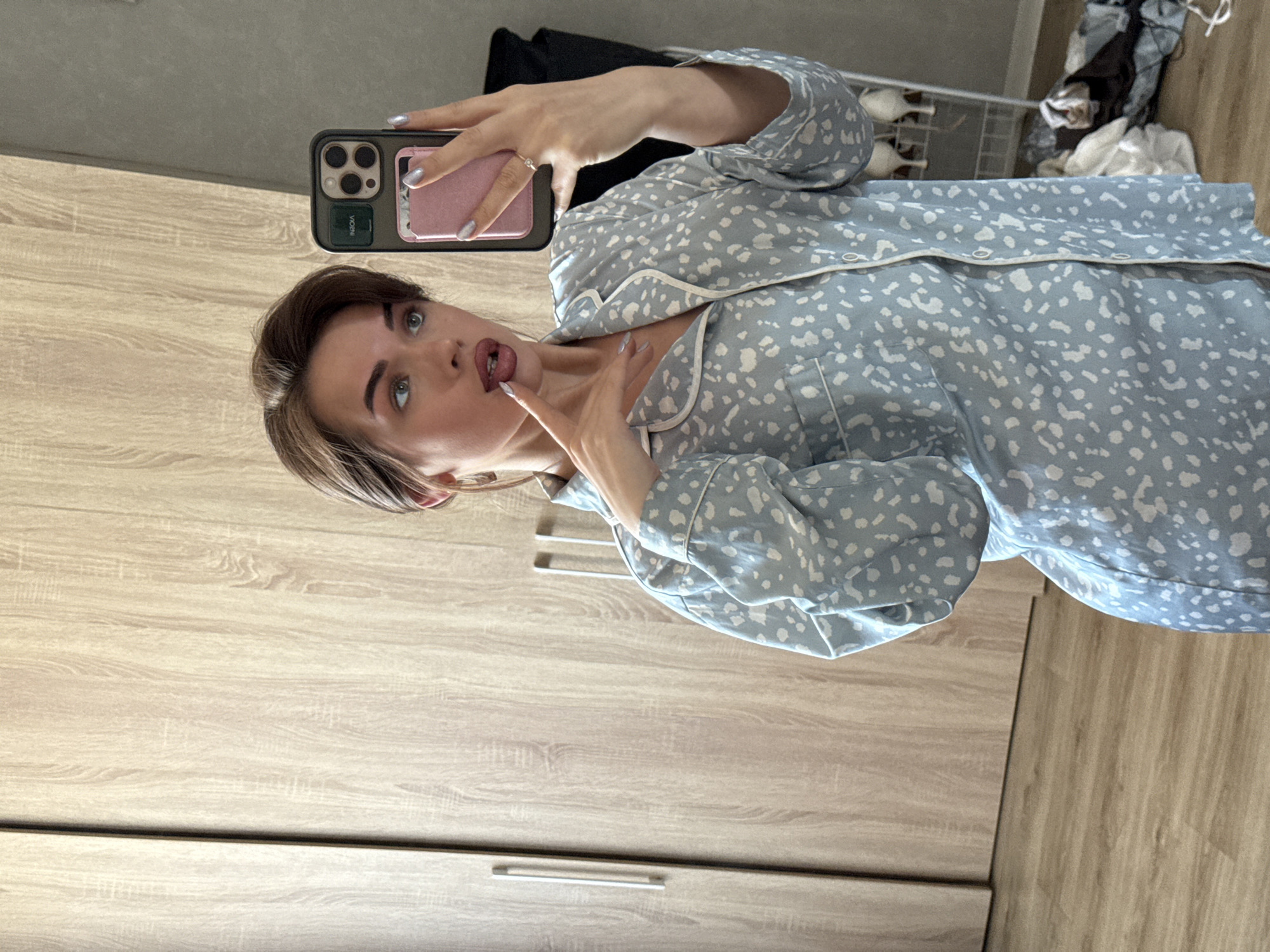 A woman is taking a selfie in a room with wooden furniture. She is wearing light blue pajamas with white spots and holding her phone in her left hand. The phone has a black and pink case. Her right hand is near her face. A metal shelf with various objects is visible in the background. The photograph was taken indoors. The location, Лобаново, Russia, is not identifiable from the image.