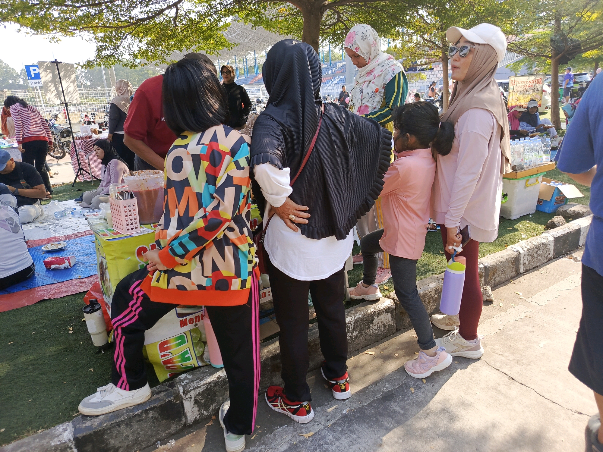 The image depicts an outdoor market scene in Tangerang, Indonesia. Several individuals are present, some appearing to be shoppers and others vendors. In the foreground, a child wearing a colorful jacket and black pants with pink stripes is visible, along with a woman in black attire and a headscarf. To the right, another child in a pink shirt and dark pants stands next to a woman wearing a light pink top, a hijab, and a white baseball cap. They are standing near a stall with bottled drinks. In the background, there are more people, market stalls with goods displayed, and a parking sign. The environment suggests a sunny day, with trees casting shadows on the ground. The overall scene captures a casual gathering and commercial activity in a public space.