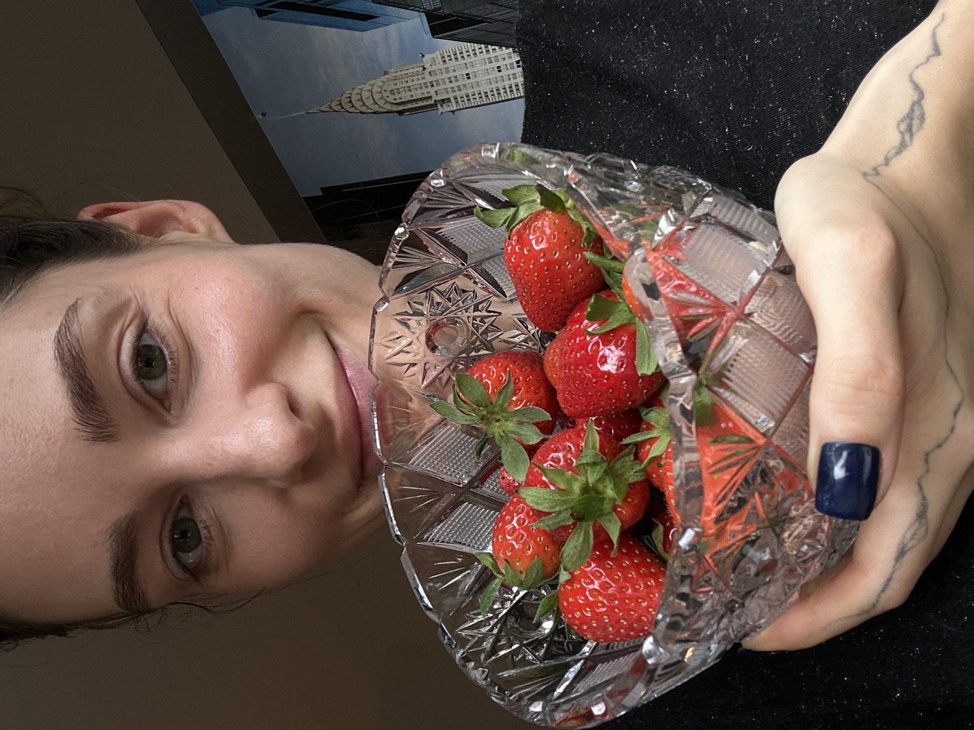 A person is holding a glass bowl of strawberries. The person is a fair-skinned individual with brown hair and green eyes. They are looking at the camera with a slight smile. Their fingernail is painted a dark blue. There is a tattoo on their hand. The bowl contains several red strawberries with green stems. In the background is a partial view of a building. It is daytime. The location in the city of Kiel, Germany is not visible in the image.