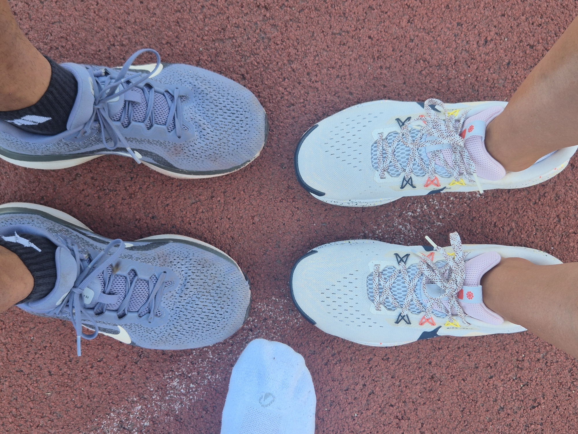 The image shows a top-down view of four feet wearing athletic shoes on a red running track. Two pairs of shoes are visible: one pair is light blue with white accents and laces, and the other pair is predominantly white with pink accents and multicolored laces. The left side of the image features two light blue shoes, one with a dark grey sock and visible skin, and the other with a dark grey sock and skin. The right side of the image shows two white shoes, one with a pink ankle sock and visible skin, and the other with a pink ankle sock and skin. A single white sock is positioned in the center of the image, between the two pairs of shoes. The scene appears to be outdoors, as indicated by the textured red running track surface and the absence of indoor elements. The lighting suggests it could be daytime. The overall impression is of individuals preparing for or having just completed a running activity. Some faint white speckles are visible on the track, possibly chalk or dust. There is no visible text in the image. The setting suggests a sports facility, likely a track and field.