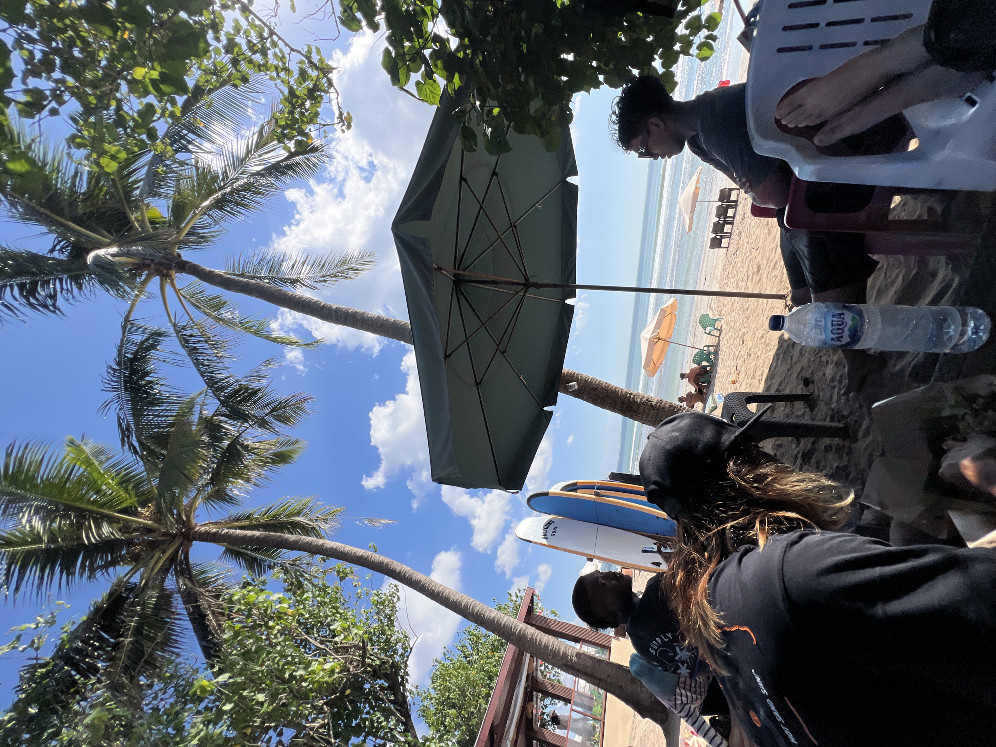 On a sunny day in Kuta, Indonesia, a group of people are relaxing on the beach under palm trees. In the foreground, three people are seated, two of them partially obscured. One person, wearing a black t-shirt and a black cap, is facing away from the camera, with their long, wavy brown hair visible. Another person, in a black t-shirt, is looking to their left. The third person, wearing sunglasses and a dark t-shirt, is seated and looking to their right, with their arms crossed. A bottle of Aqua water is placed on the sand beside them. Behind them, several surfboards stand upright. In the background, the ocean is visible with gentle waves, and beach umbrellas dot the sand. Palm trees with lush green fronds dominate the upper portion of the image, with a clear blue sky dotted with white clouds above. A shaded, open-air structure with a red roof is visible on the left side of the frame. The overall atmosphere suggests a relaxed beach day in a tropical setting.