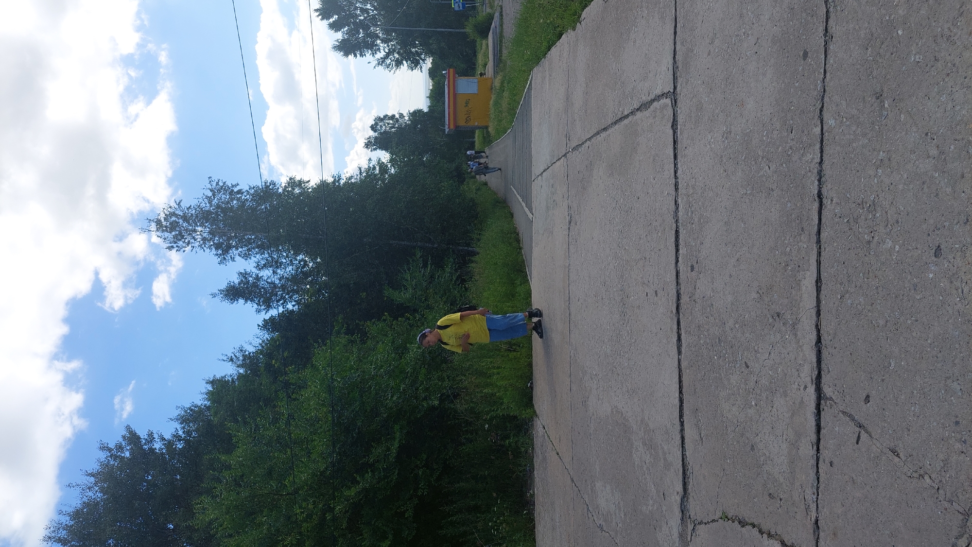 In Amursk, Russia, a young person with a yellow t-shirt and blue pants walks on a concrete pathway. The pathway is cracked and weathered, bordered by lush green trees and tall grass. A yellow bus stop shelter with a blue-tinted window stands further down the path, with several people walking in the distance towards it. The sky is a bright blue with scattered white clouds, and the overall scene suggests an outdoor, possibly rural or suburban, setting.