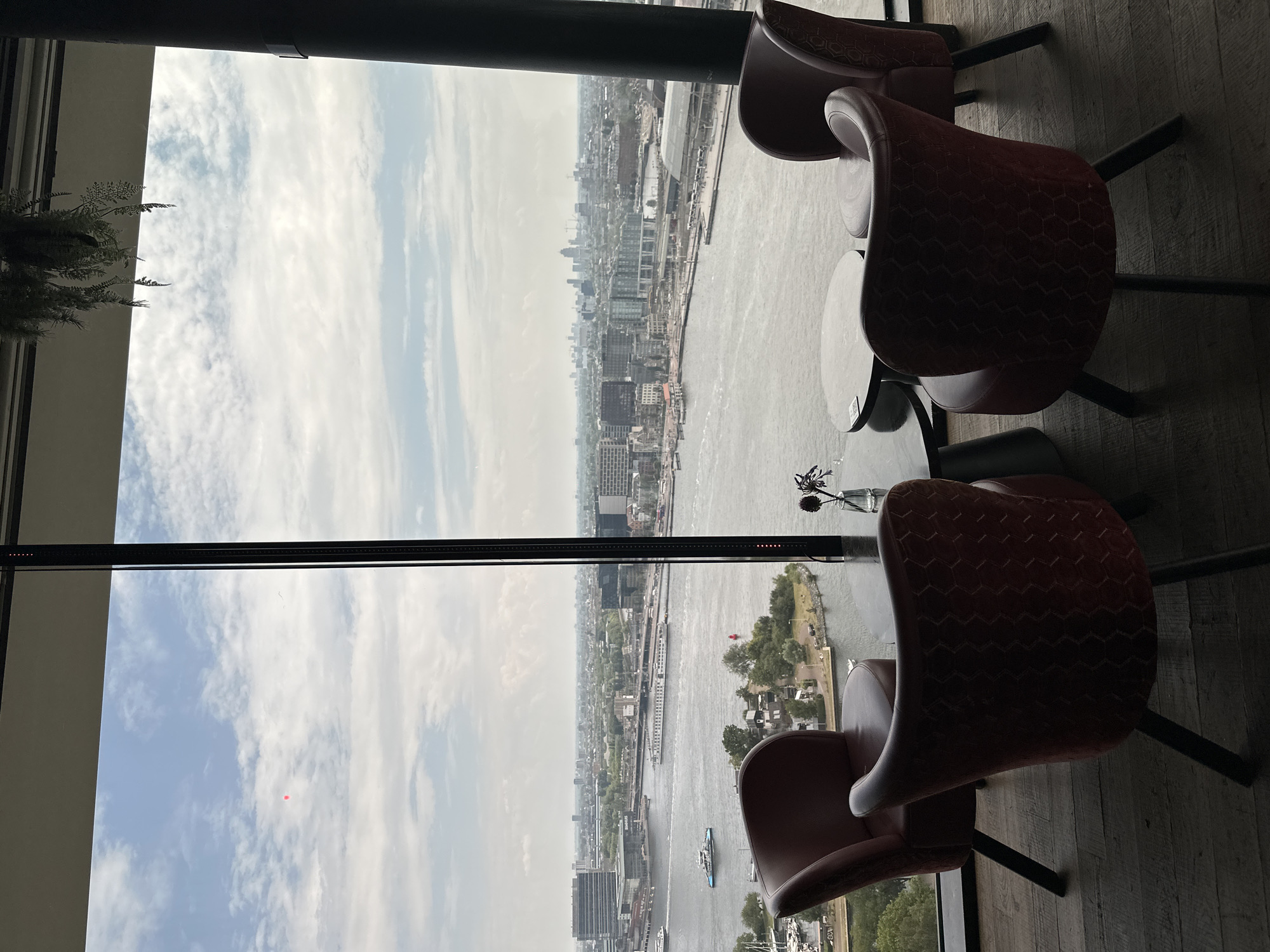 The image depicts a panoramic view of Amsterdam, Netherlands, from a high vantage point, likely a restaurant or lounge. In the foreground, four red upholstered chairs with dark legs surround a round, dark-colored coffee table. A small glass vase with a single, dark purple flower sits on the table. The chairs are positioned to face the expansive window overlooking the city and a wide body of water, possibly a canal or river.
Beyond the seating area, the scene transitions to a detailed urban landscape. The water is dotted with various vessels, including ferries and what appear to be tour boats. Buildings of different architectural styles and heights line the waterfront and extend into the distance, forming the characteristic Amsterdam skyline. Notably, a large, modern building with a curved facade is visible on the left, and further in the distance, more contemporary structures and construction cranes punctuate the horizon. The sky above is partly cloudy, with a mix of white cumulus clouds and patches of blue. A faint red object, possibly a balloon or drone, is visible in the upper left quadrant of the sky. Lush greenery, trees, and smaller buildings are scattered along the edges of the waterway, suggesting a blend of urban development and natural elements. The overall impression is one of a sophisticated urban setting offering a commanding view of the city.