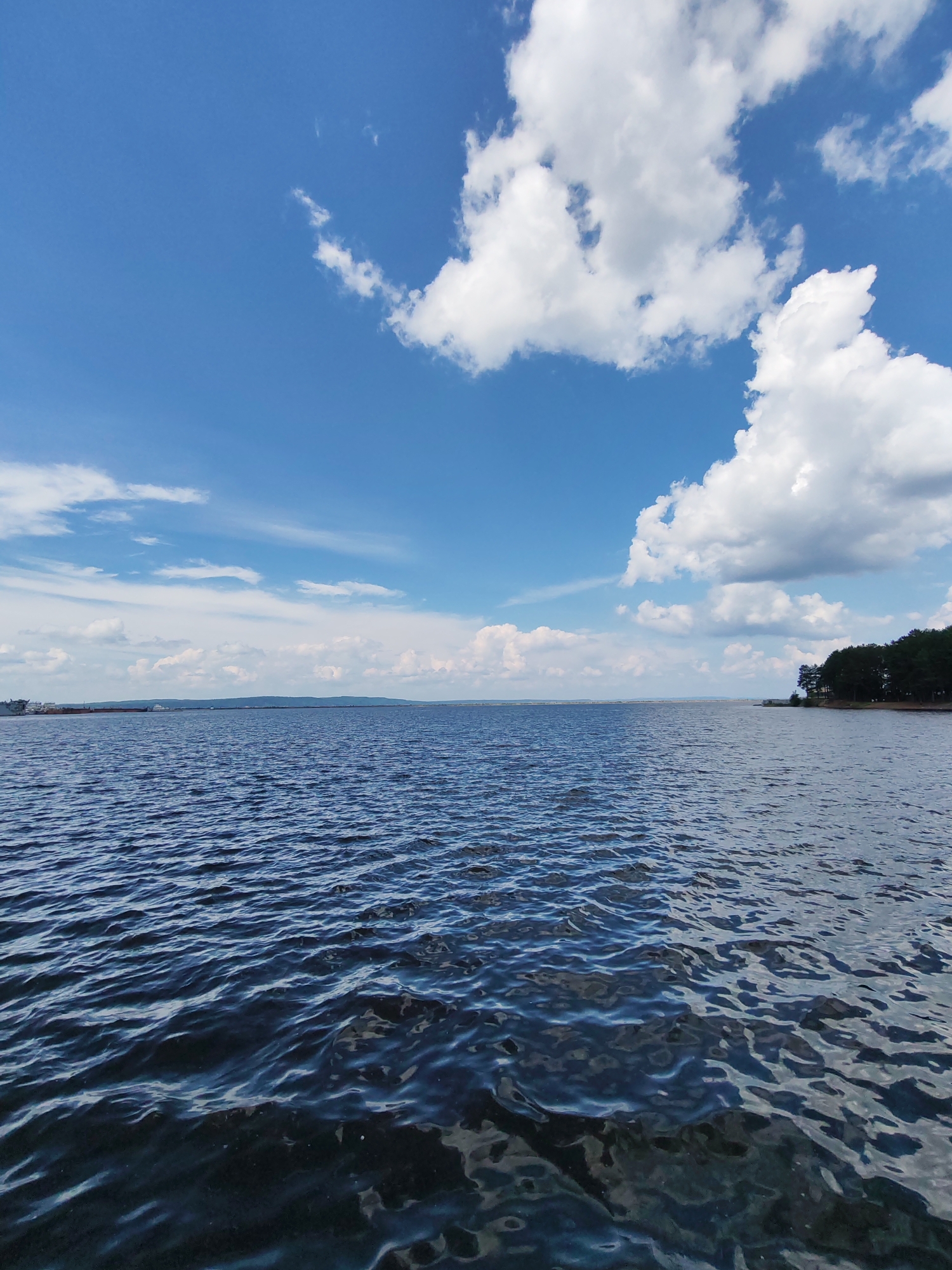 A wide-angle, eye-level outdoor shot captures a tranquil body of water under a partly cloudy blue sky. The foreground is dominated by the dark blue surface of the water, with small, gentle ripples reflecting the sky's light. In the mid-ground, the expanse of the water stretches towards a distant, hazy shoreline. Along the horizon, a faint line of land is visible, punctuated by what appear to be buildings and a line of trees on the right side of the frame. Large, fluffy white clouds are scattered across the bright blue sky, with a few wispy clouds trailing in the distance. The overall scene suggests a peaceful day by the water. This view is likely of the Kama River in Chaikovsky, Russia, a prominent feature of the city known for its industrial and natural beauty. No people or specific actions are visible in the image.