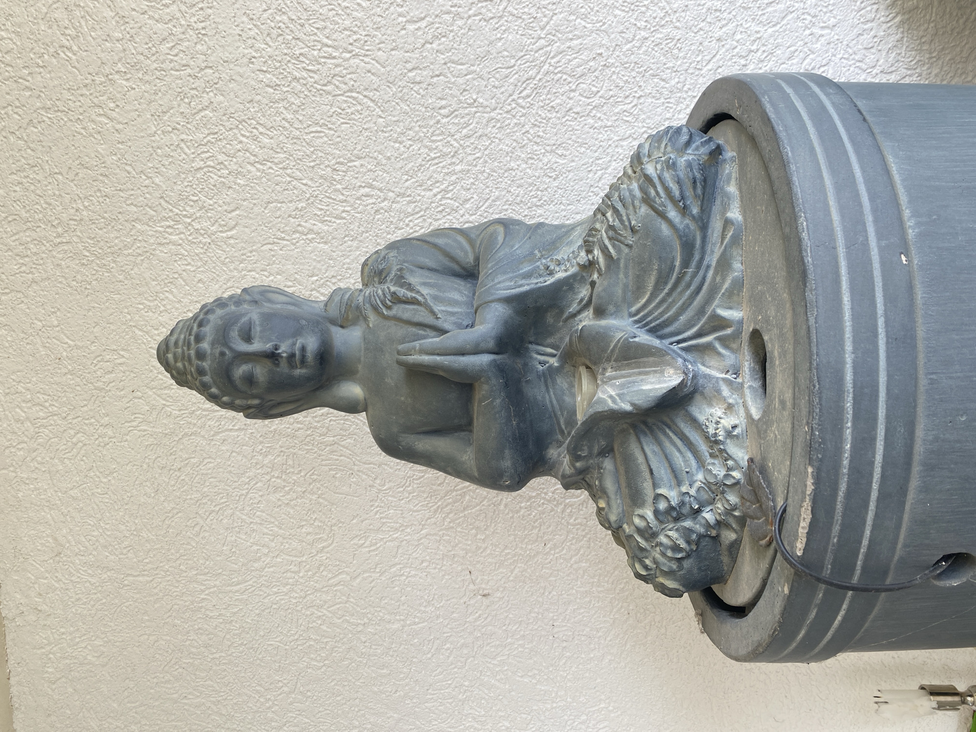 A stone Buddha statue is affixed to a round, grey, horizontal object with ridges, possibly a decorative fountain element, set against a textured white wall. The image appears to be taken indoors. There are no people visible. There is no location in the city of Trebesin, Montenegro.