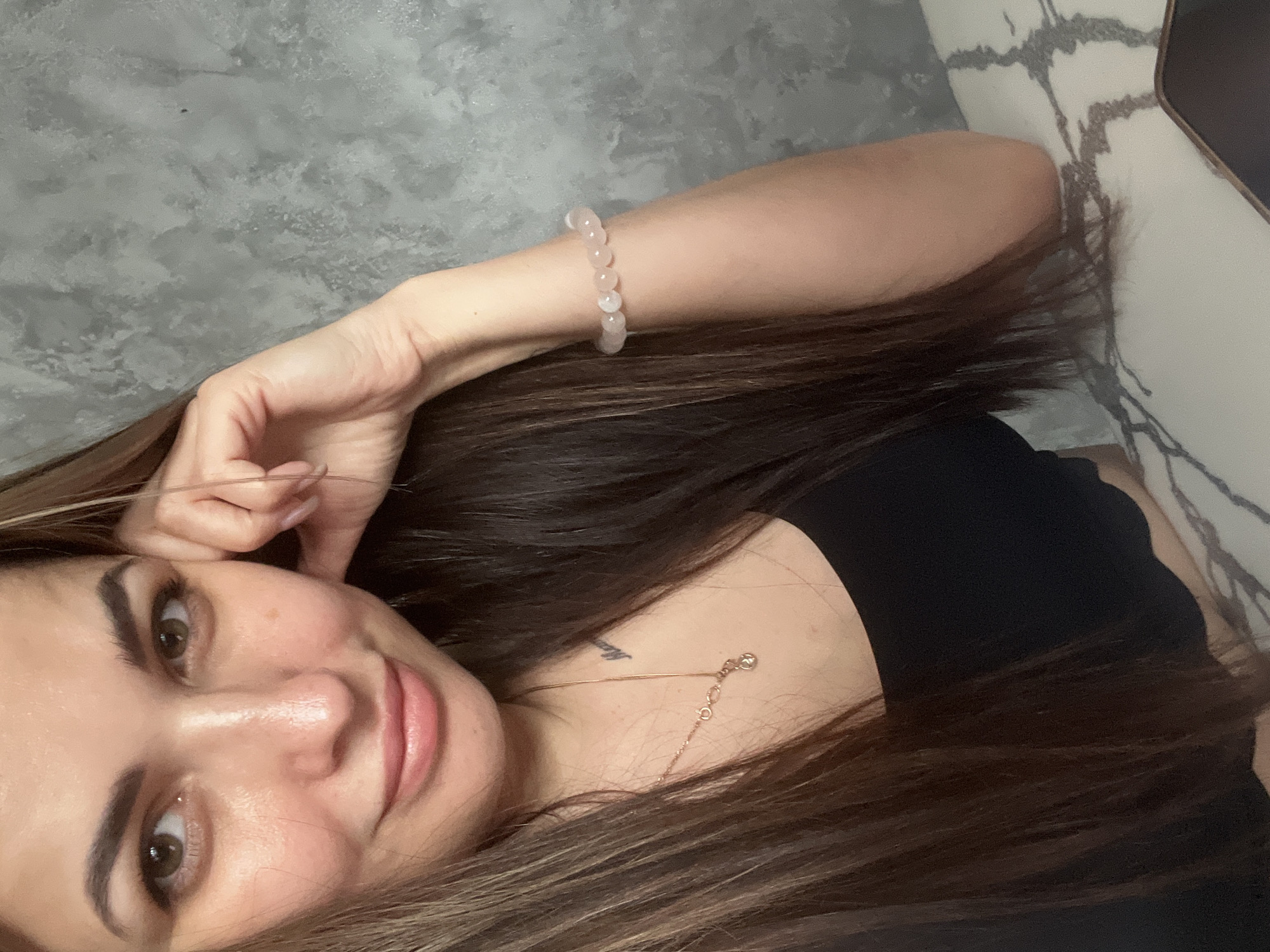A woman is photographed indoors. Her brown hair is long and wavy. She is wearing a black top and a bracelet made of round, light pink beads. A gold necklace is also visible. The background consists of a gray wall and a white patterned surface. The photograph appears to be taken in Unknown, Unknown.