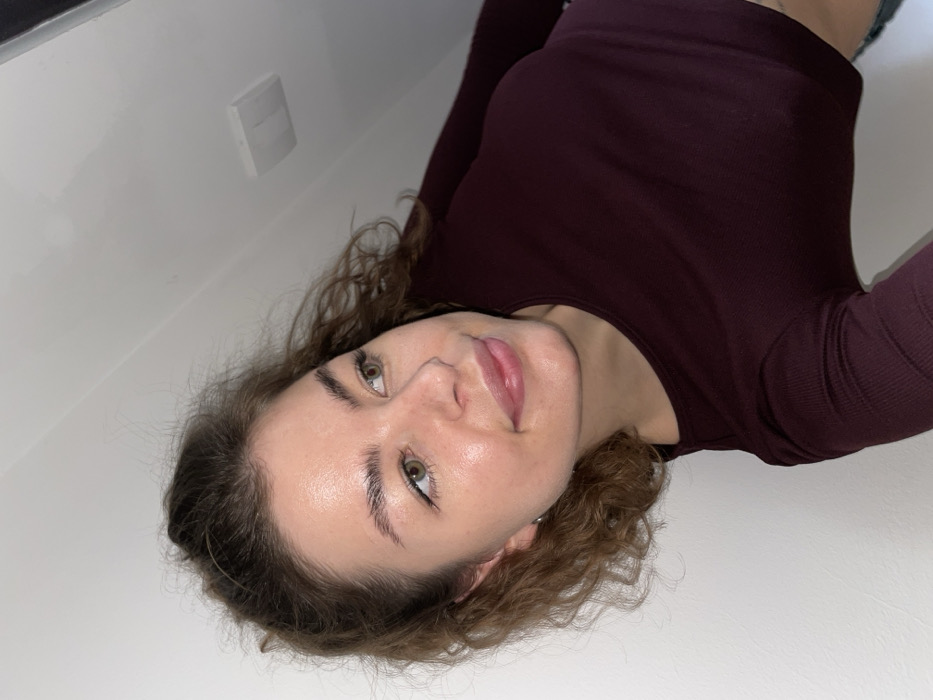 The image displays a close-up, tilted portrait of a single female subject within an indoor environment. The precise location in Boreti, Montenegro, is not discernible from the visual information provided. The subject is a young adult with light skin, medium-length curly brown hair pulled back, and green or hazel eyes. Her eyebrows are well-defined, and her lips appear full. She exhibits a slight, closed-mouth smile and is gazing towards the viewer's right. She is attired in a long-sleeved, ribbed, dark maroon or burgundy crop top. A small, circular silver earring is visible on her left earlobe. The background consists of a plain white wall and a white ceiling, where a single white rectangular fixture, likely a light cover or ventilation unit, is mounted above and to the right of the subject's head. The scene is brightly illuminated, consistent with artificial lighting.