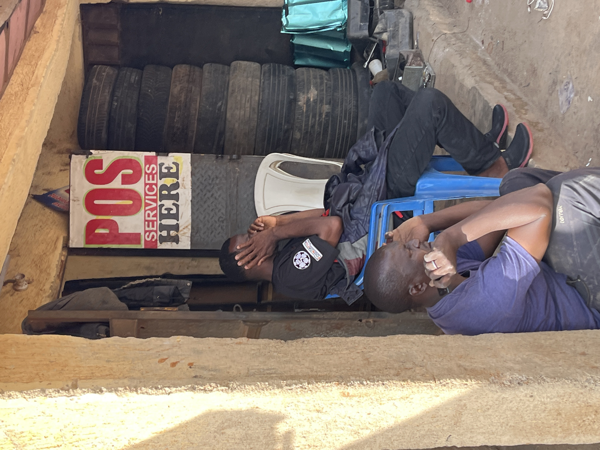 In Barakin Akawo, Nigeria, an outdoor stall displays "POS SERVICES HERE" on a red and white sign. Two men are present. One man, wearing a black t-shirt with a circular logo and dark pants, sits on a blue plastic chair, covering his face with his hands. The other man, in a purple t-shirt and dark pants, sits on the ground to the left, leaning against a rough, beige-colored pillar, also with his hand near his face. Behind the seated men, a stack of tires is visible, along with turquoise bags and various tools and electronic equipment. The ground is dirt and debris.