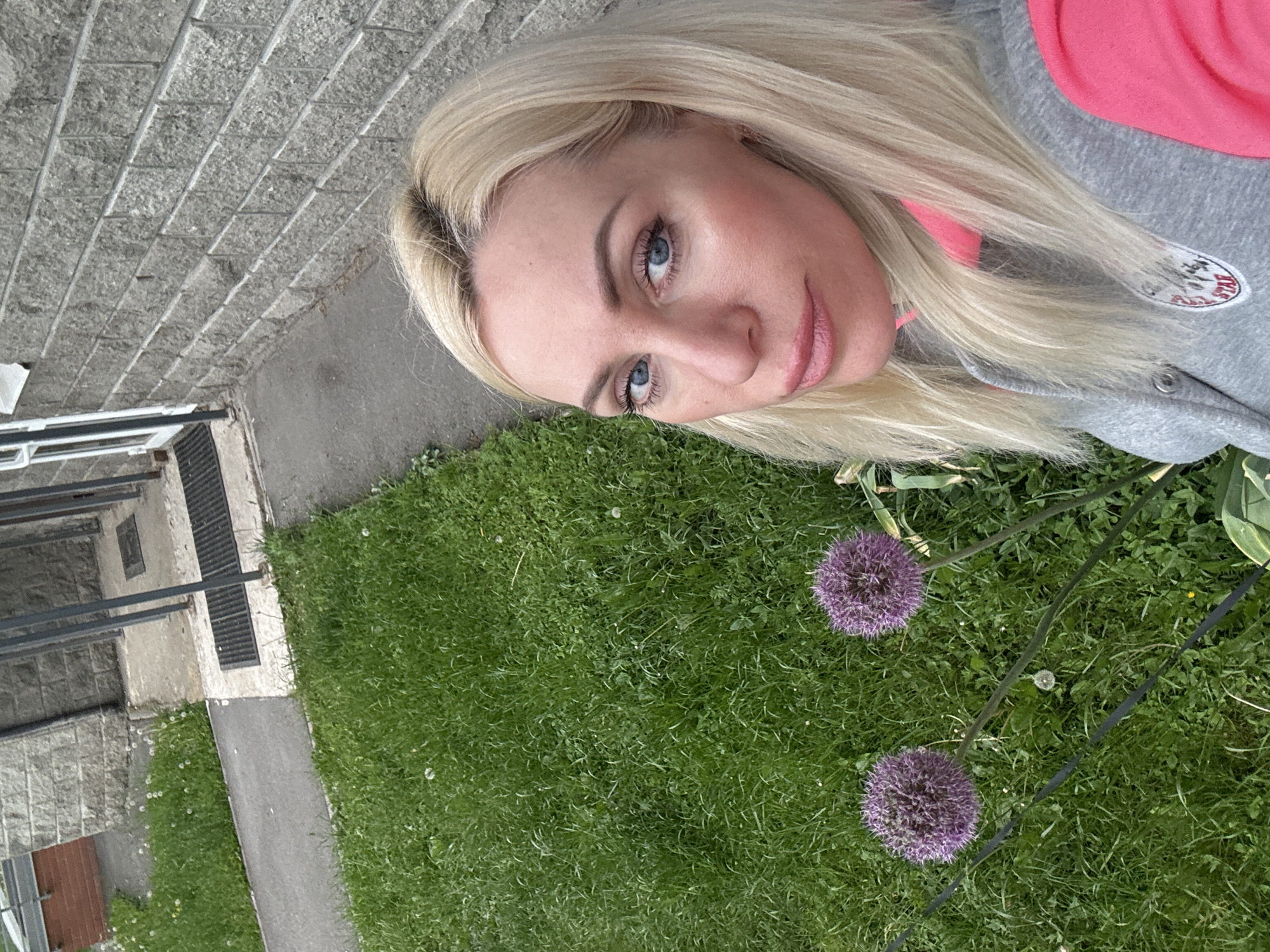 A woman with blonde hair and blue eyes is posing for a selfie outdoors. She is wearing a gray and pink hooded sweatshirt. Behind her is a patch of green grass with two purple allium flowers. In the background, there is a brick wall and a metal grate. The location appears to be an urban area.