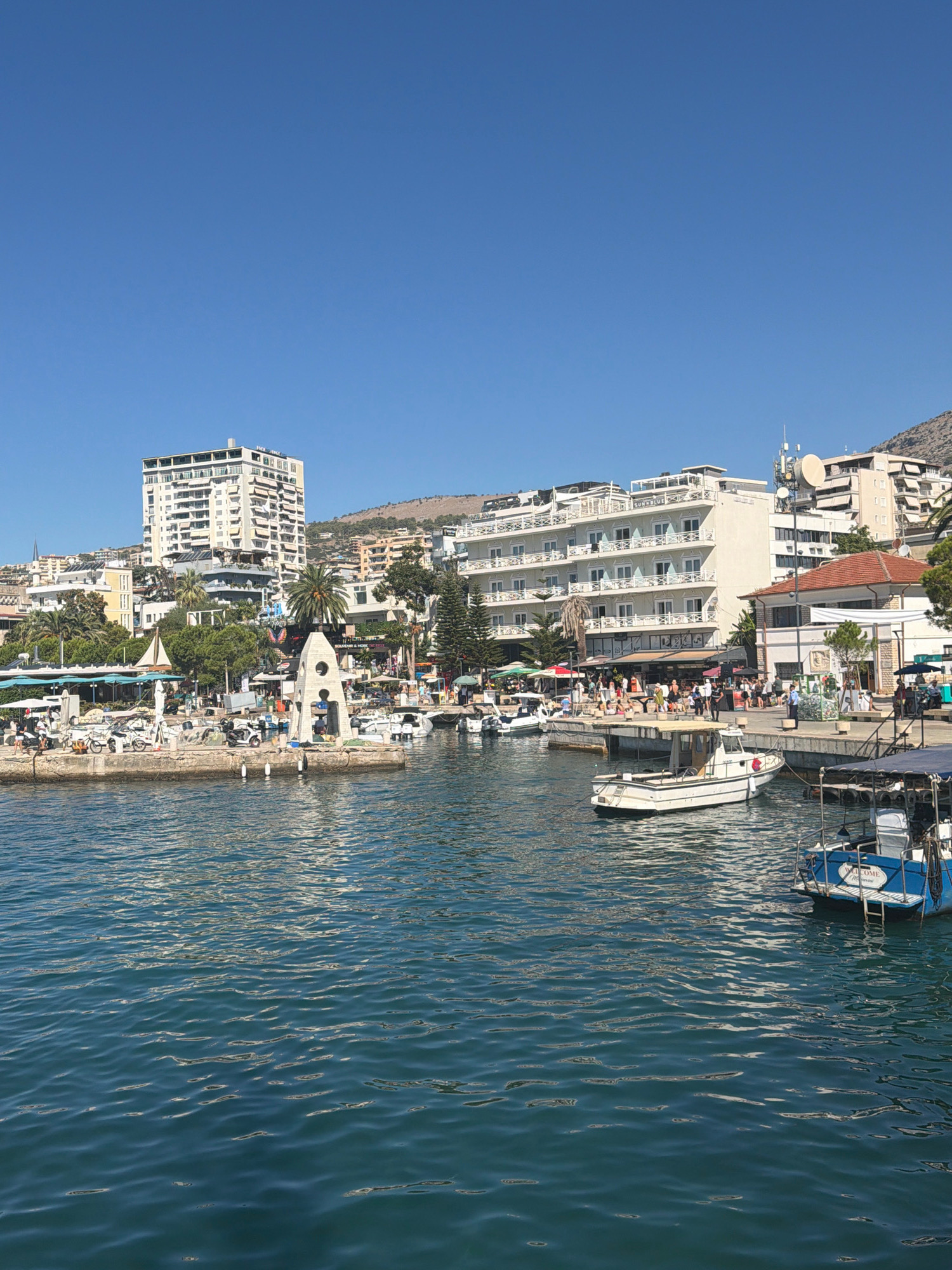 This is a daytime, eye-level shot of the Saranda, Albania waterfront. The scene is a harbor with several boats docked and moored. In the foreground, the deep blue water of the harbor is calm with slight ripples. A blue and white boat is docked to the right, with the word "WELCOME" visible on its stern. Behind it, a white boat with a cabin is also docked.
The middle ground features a stone pier extending into the water. Along the pier and the adjacent boardwalk, there are various people walking and standing. Several shops and restaurants with outdoor seating areas are visible along the waterfront, adorned with umbrellas and awnings. A distinctive white, conical monument with an arched opening stands on the pier. Scooters and bicycles are parked on the pier.
In the background, a row of multi-story buildings lines the coastline. The buildings are a mix of modern hotels and older structures, many with balconies and light-colored facades. A tall, white building with many balconies stands prominently. Further up the hillside, more buildings and vegetation are visible. The sky is a clear, bright blue. The overall impression is a bustling, sunny day at a popular coastal resort.