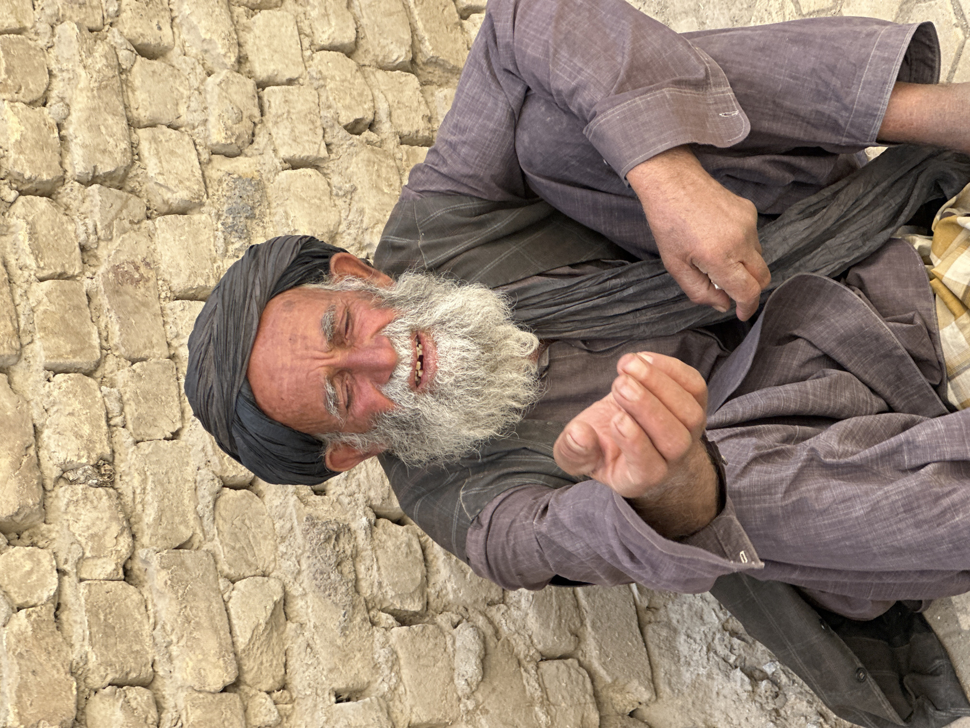 An elderly man with a white beard and a dark turban sits against a brick wall in Kanozai, Pakistan. His eyes are closed and his mouth is open, revealing broken teeth. He is wearing a grey shirt and pants, with a dark scarf around his neck. His right hand is outstretched, palm up, as if begging. The brick wall behind him is made of rough, irregular stones.