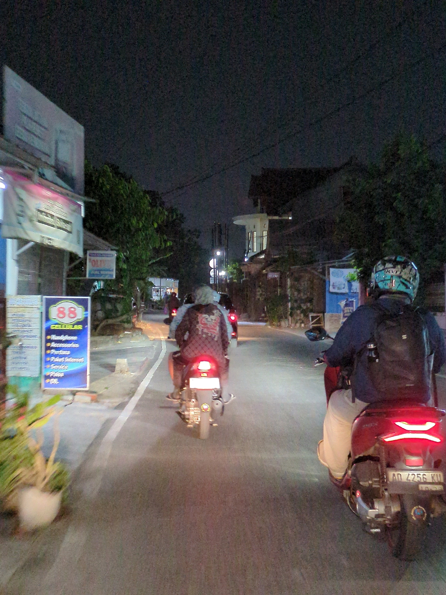 The image depicts a nighttime scene on a street in Ngadirejo, Indonesia, likely during a period of low visibility due to the darkness. The perspective is from behind a person riding a red scooter, who is facing away from the camera. The scooter has a license plate that reads "AD 4256 KU." The rider is wearing light-colored pants and a dark shirt, with a black backpack. They are in motion, indicated by the blur of the road and the motion of other vehicles.
In front of the viewer's scooter, another person is riding a scooter, also facing away from the camera. This rider is wearing a patterned jacket and a helmet. Further ahead, more scooters are visible on the road, some with their brake lights illuminated.
The environment includes buildings and trees lining the street. On the left side of the road, there are several signs, including a large, brightly lit sign with the number "88" and the word "CELULAR" below it, listing items like "Handphone," "Accessories," and "Paket Internet." There are also potted plants visible near this sign. The road surface is asphalt with white lane markings. The overall lighting is dim, with artificial lights from streetlamps and vehicle headlights providing illumination. The sky is dark and overcast, suggesting a clear night or the presence of cloud cover. The image appears to be taken from another moving vehicle, as evidenced by the motion blur.