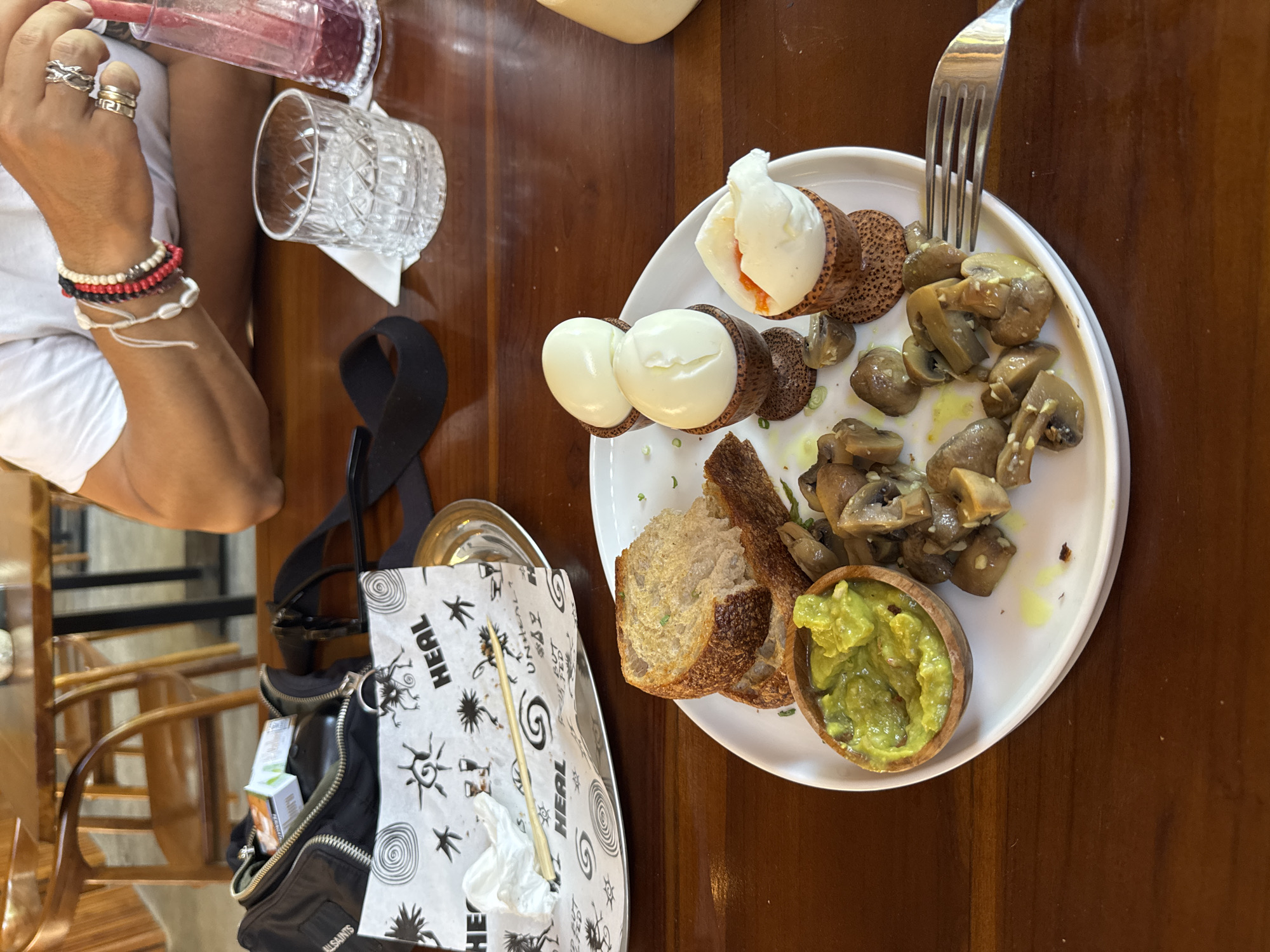 The image depicts a dining scene on a dark wooden table, likely in a restaurant in Kuta Selatan, Indonesia. A plate of food occupies the center of the frame, containing sliced eggs served within wooden casings, sauteed mushrooms, slices of bread, and a wooden bowl of guacamole. A fork is partially inserted into the mushrooms. To the left of the plate is a metal tray lined with patterned paper, partially obscured by a black bag with visible items inside. Further left, a person is visible from the shoulders up, wearing a white shirt, bracelets, and rings. A glass of clear liquid and a taller glass with a pinkish liquid are positioned near the person. The scene is well-lit, and the environment appears to be an indoor dining area.