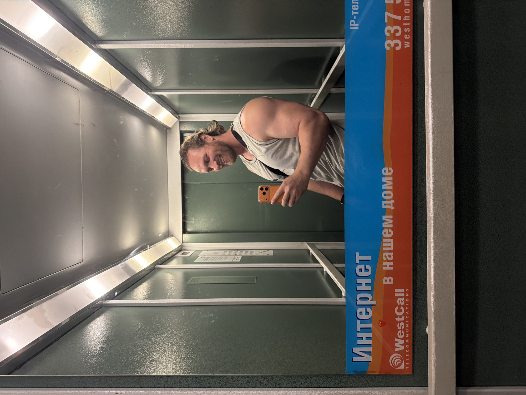 A man is taking a mirror selfie in an elevator. He is a white male with blonde hair tied back in a ponytail and a beard, wearing a grey tank top. He is smiling and holding an orange smartphone in his right hand. The elevator has green walls and is lit by overhead fluorescent lights. There is an advertisement panel at the bottom of the image with Russian text that reads "Интернет в нашем доме" which translates to "Internet in our home", and a logo for "WestCall TELECOMMUNICATIONS". The man is in an elevator in Saint Petersburg, Russia.