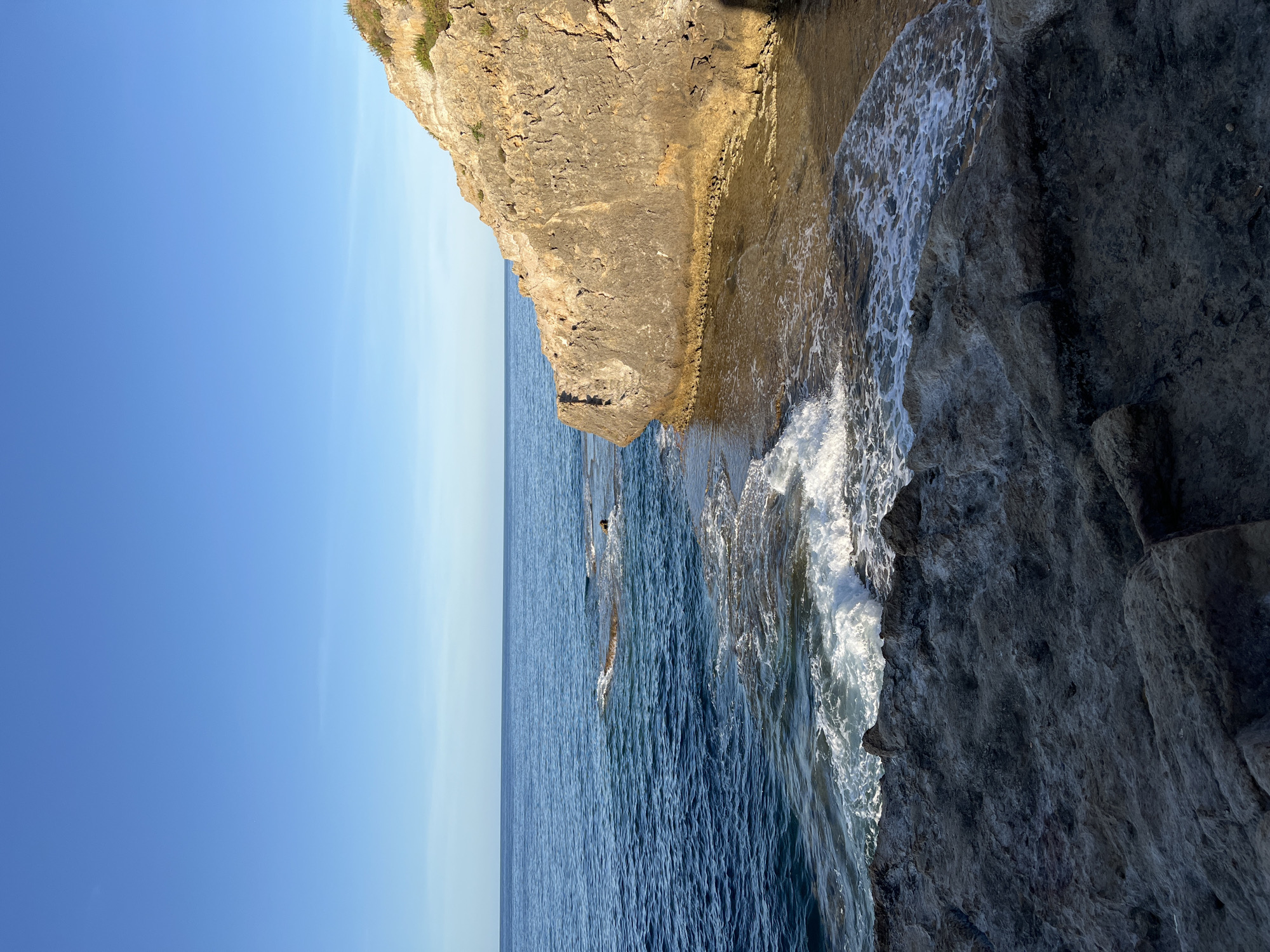 The image depicts a rocky coastal scene in Sperone, Italy, under a clear, transitioning blue sky. The upper portion of the frame features a cloudless sky, light blue at the horizon and deepening overhead. Below, the calm, deep blue sea extends to the horizon, with gentle waves breaking on the foreground and midground rocks. In the foreground, dark, textured, irregular rock formations occupy the lower left and central areas. To the right, a prominent, light brown, stratified cliff face rises, adorned with sparse green vegetation on its upper edge. The base of this cliff meets the water, forming a shallow, light-colored inlet where gentle ripples are visible. No individuals, animals, or man-made objects are present in the scene.