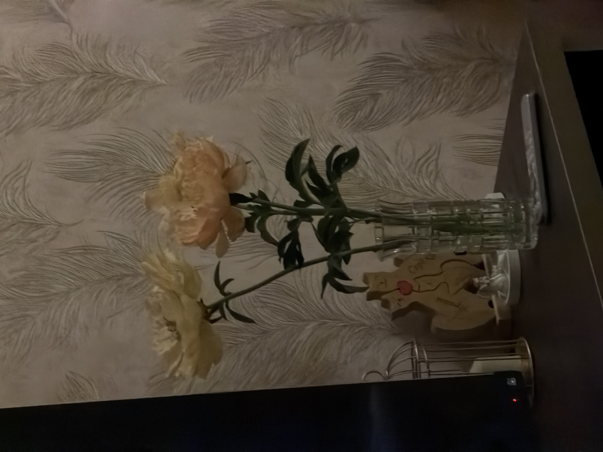 The image displays an indoor still life scene, rotated 90 degrees clockwise from its natural orientation, featuring decorative objects on a dark surface against patterned wallpaper.
In the center, two large, light-colored flowers, likely peonies or similar blooms in a cream or pale peach hue, with green stems and leaves, are arranged in a clear, vertically ribbed glass vase. The vase stands on a dark, flat surface, which appears to be a shelf or tabletop.
To the right of the vase, a wooden decorative item or puzzle is visible. It is intricately cut, resembling a stylized human torso or an abstract shape, and features a prominent red heart-like detail near its center. Above this red detail, the Cyrillic word "Сердце" (Russian for "Heart") is clearly visible. Further to the right, a small metallic object, possibly a cage-like lantern or candle holder, is partially seen. Below these items, a dark rectangular electronic device with a small red indicator light is present.
The background consists of a light-colored wall adorned with wallpaper featuring a repeating pattern of delicate, swirling feather or leaf-like motifs in a slightly contrasting tone.
The scene is illuminated by dim, warm lighting, suggesting an evening or interior setting. The overall atmosphere is calm and decorative. The location context provided is Izhevsk, Russia.