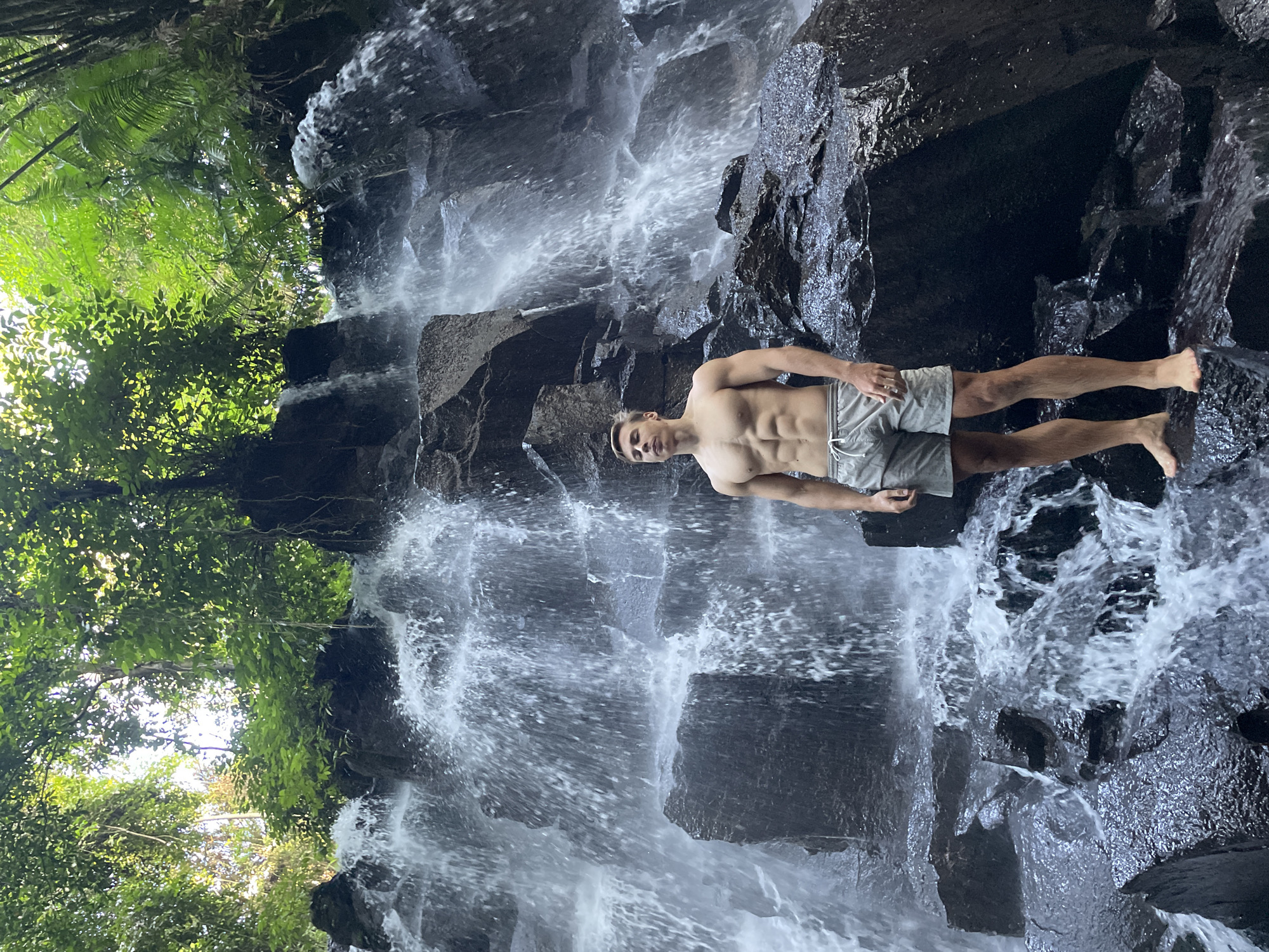 A shirtless man in gray shorts stands on a dark rock formation in front of a waterfall. The waterfall is divided into several streams cascading down the rock face. To the left of the waterfall, green foliage is visible. The background and foreground consist of rock and flowing water. The photo seems to have been taken at the Tegenungan Waterfall near the city of Gianyar, Indonesia.