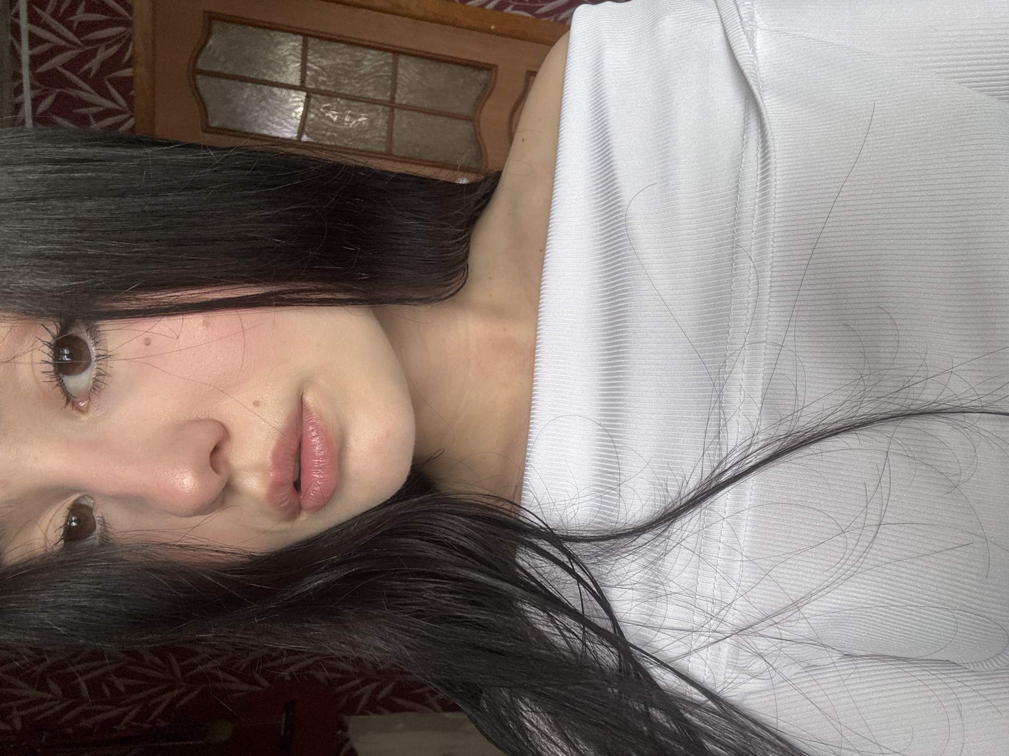 A close-up, rotated image features a person with dark hair and light skin. Part of their face is visible, including eyes, nose, and lips, and they appear to be wearing a white top. The background shows a wooden door with decorative panels and a patterned red surface.
The geographical location in Фоки, Russia is not discernible from the image.