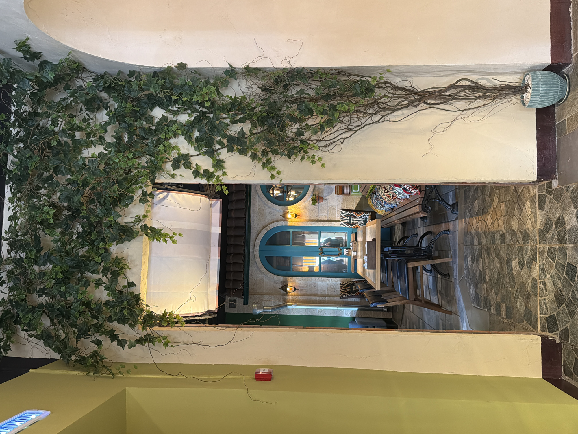 This image presents a view into an empty, warmly lit dining or cafe area, characterized by its Mediterranean-inspired or rustic decor. The perspective is from an entry point, looking through an opening into the main seating space.
A significant portion of the foreground on the left is dominated by a wall extensively covered with artificial ivy and intertwined bare, gnarled branches. This greenery originates from a decorative light blue-green pot, filled with small white stones, positioned on the far right end of this wall.
Beyond the initial opening, the main dining area unfolds. Its central feature is a distinctive arched double door, painted a vibrant teal color, with glass panels. Lantern-style wall sconces, emitting a warm glow, flank this unique entrance. Inside, several wooden tables and chairs are arranged, some fitted with colorful, patterned cushions or throws. The floor of this area is paved with gray and black tiles, laid out in an intricate, circular pattern. The walls in the dining section have a textured finish, resembling stone or rough plaster.
In the lower foreground, a plain light-green wall section is visible, hosting a small red box, possibly a fire alarm or electrical unit. A partial blue sign with white text, appearing to read "OYAS" or similar, is faintly visible at the very bottom left.
The scene is illuminated by artificial light sources, creating a cozy and inviting ambiance. There are no people visible, suggesting the establishment might be closed, or it's a quiet time of day. The combination of artificial foliage, unique architectural elements, and patterned soft furnishings contributes to a thoughtfully designed and welcoming atmosphere.