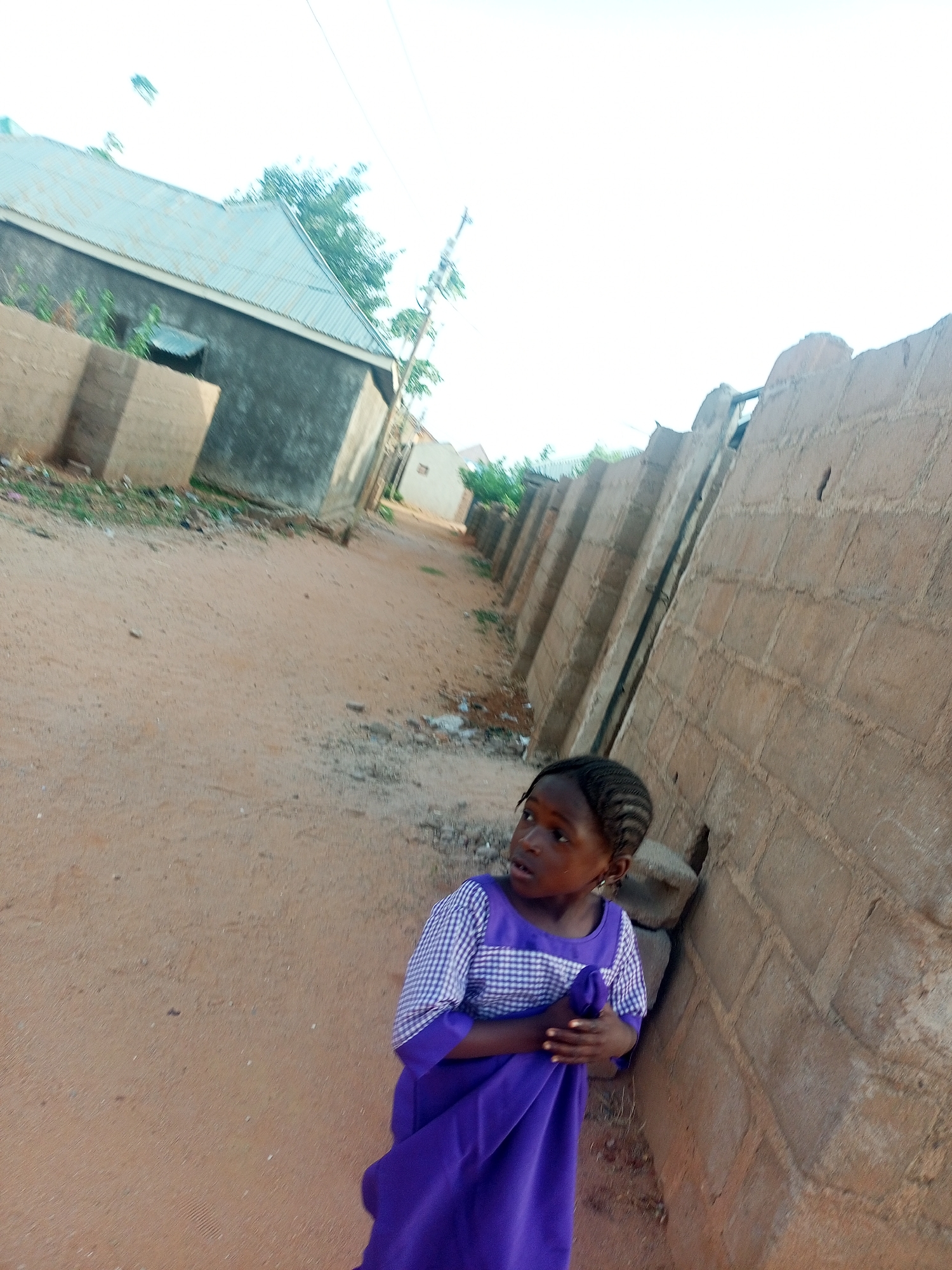 A young girl in a purple and white dress stands next to a brick wall in a dusty, undeveloped area. The girl looks off to the left with a concerned expression. In the background, there are buildings with corrugated metal roofs. The ground is dirt and sand. The scene appears to be a street or alleyway in Tempure, Nigeria.