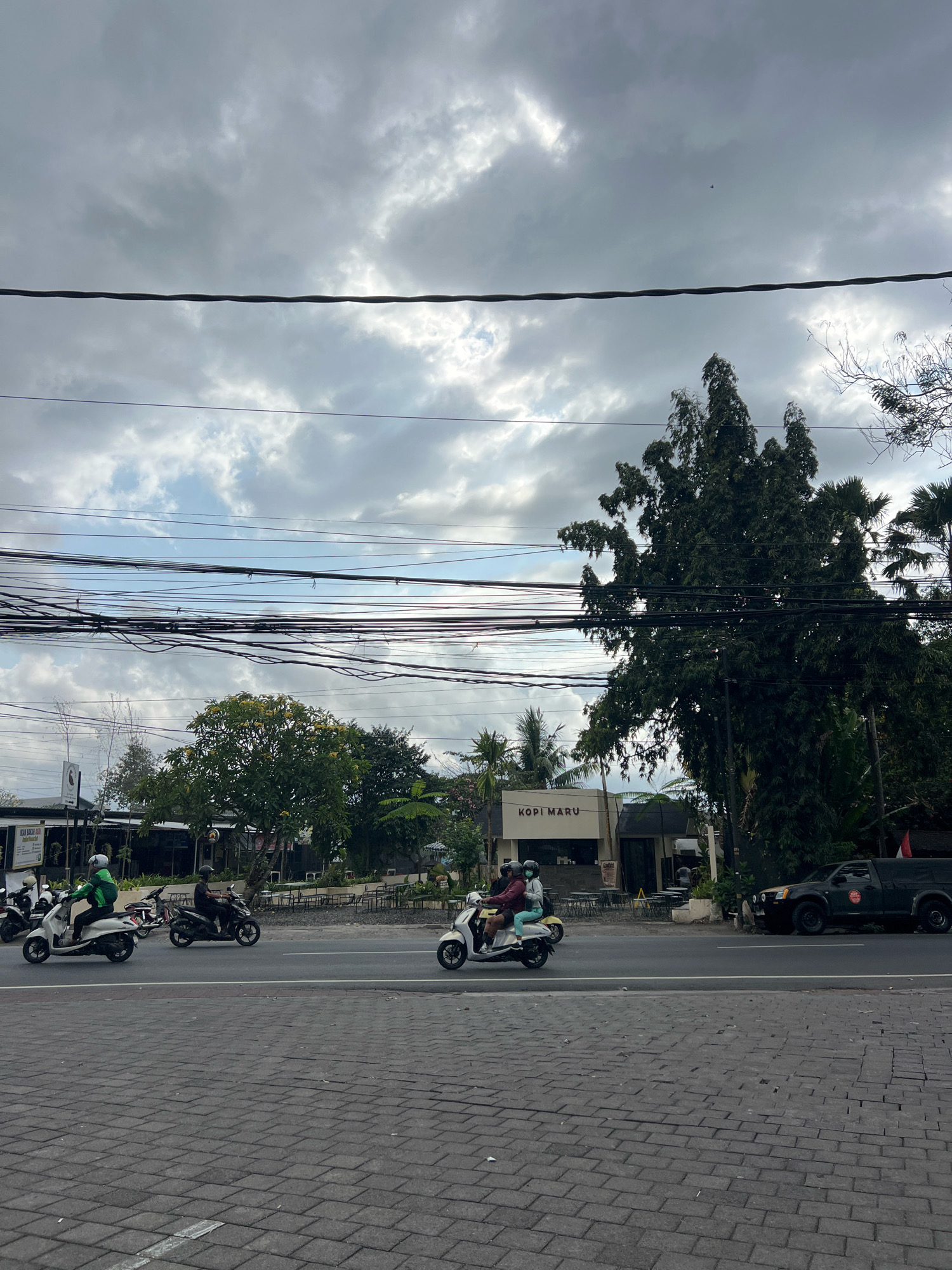 The image captures a street scene in Denpasar, Indonesia, under a cloudy sky. In the foreground, a brick-paved sidewalk runs along a road where several scooters are in motion. On the road, three scooters are visible, each carrying one or two passengers, all wearing helmets. The first scooter, on the left, is white and ridden by a person wearing a green jacket and a white helmet. The second scooter, dark in color, has a single rider with a helmet. The third scooter, also white, carries two passengers, one in a maroon jacket and the other in a light blue top, both wearing helmets. Further down the road, a dark-colored pickup truck is parked.
Behind the road, there is a low-rise building with a sign that reads "KOPI MARU". The building appears to be a cafe or restaurant, with outdoor seating visible in front. Lush green trees and palm trees are scattered throughout the scene, some with vibrant yellow flowers. A complex network of overhead power lines crisscrosses the sky, adding a visual layer to the urban landscape. The overall atmosphere is that of a typical day in a bustling Indonesian city, with the overcast weather contributing to a subdued lighting.
