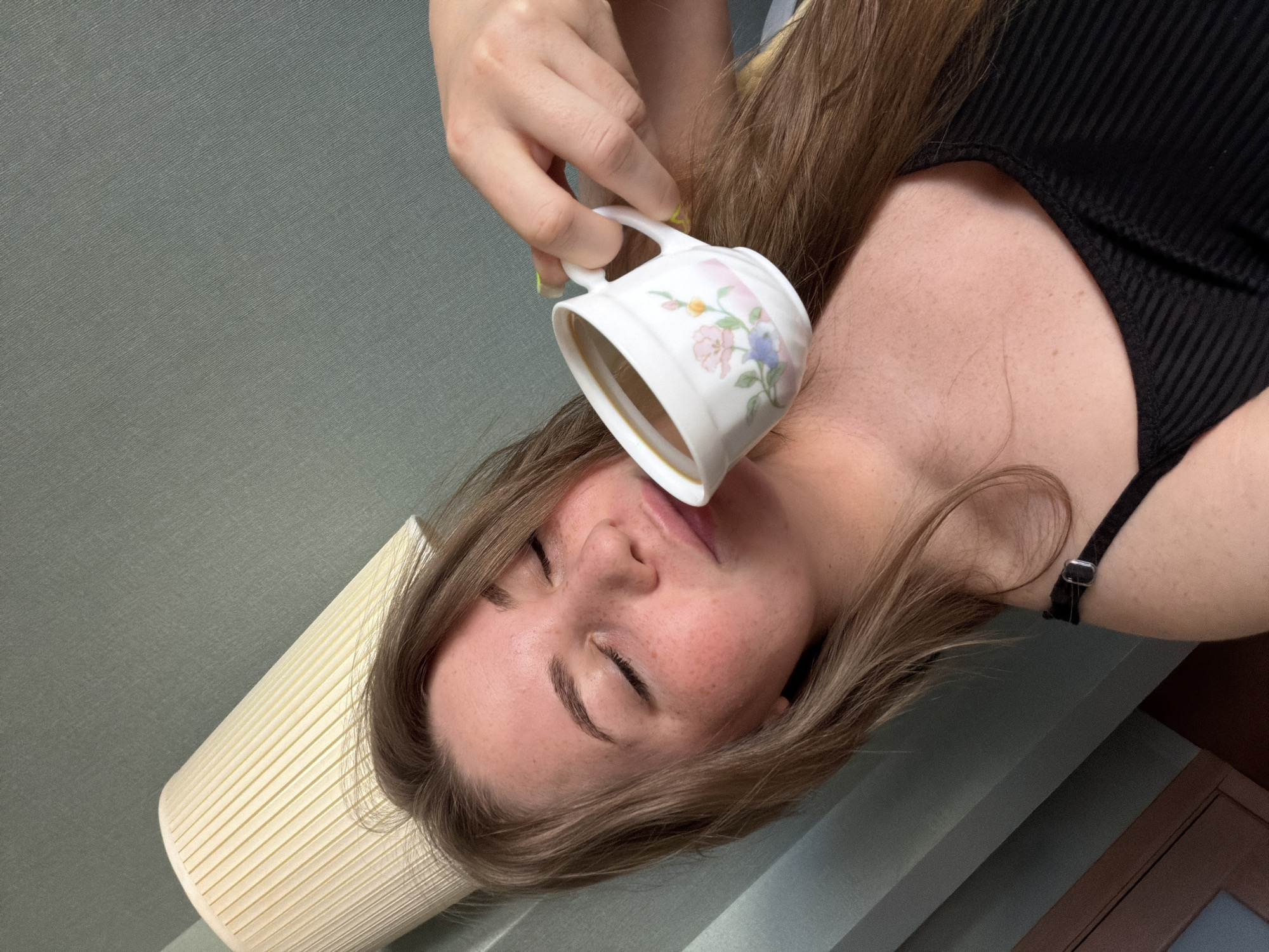 A young adult female is depicted in an indoor setting, reclining with her eyes closed and a tranquil expression. She has long, light brown hair, fair skin, and noticeable freckles across her nose and cheeks. Her lips are gently pressed to a white ceramic teacup, decorated with a floral pattern in pink, blue, and green, which contains a dark brown liquid. She is wearing a black ribbed spaghetti strap top, and her right hand, with bright yellow-green nail polish, grasps the cup. The background features a muted greenish-teal wall with a subtle fabric-like texture, and a pleated, off-white cylindrical lampshade is partially visible in the upper right. In the upper left quadrant, a dark brown surface, possibly a piece of wooden furniture or paneling, is visible. The lighting is soft and diffused.