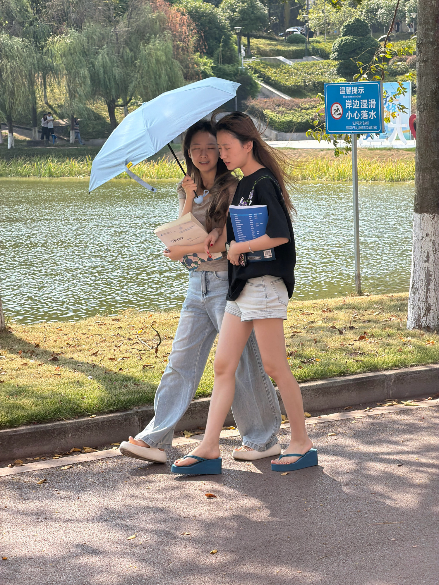 Two young women walk side-by-side on a paved path next to a body of water, likely a lake or river. The woman on the left, wearing light-wash jeans and white slip-on shoes, holds a book. The woman on the right, in denim shorts and blue wedge flip-flops, holds a different book. The woman on the left is holding a light blue umbrella open over both of them, suggesting it is either sunny or raining. They appear to be engaged in conversation, with both women smiling. Behind them, a signpost with Chinese characters and an English translation reading "Warm reminder Slippery bank Risk of falling into water" is visible, indicating a potential hazard near the water's edge. The environment suggests a park or campus setting, with trees and manicured grass surrounding the path and water. This scene is located in Jiangjin, China.