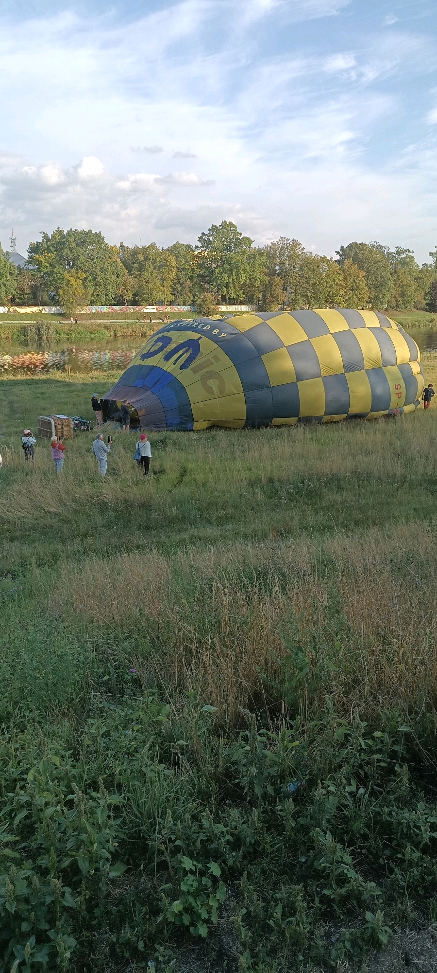 An aerial view shows a deflated hot air balloon lying on its side in a grassy field near the Oder River in Opole, Poland. The balloon is yellow and grey with a checkered pattern and has "INSPIRED BY ONIC" written on it. A group of people are gathered around the balloon, appearing to be preparing it for inflation or packing it away. In the background, trees line the riverbank, and a city skyline is visible. The sky is partly cloudy.