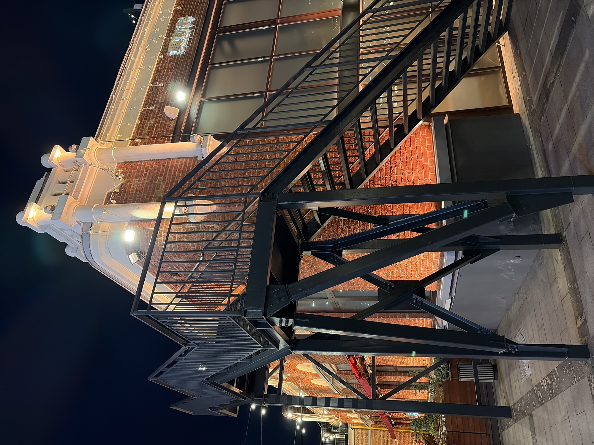 This is a night photograph taken outdoors at street level. The scene features a red brick building with white decorative elements and a black metal staircase extending upwards. The building is illuminated by artificial lights, including string lights along the facade and spotlights. The staircase has a metal railing and appears to be a fire escape or an external access route. The ground in front of the building is paved with cobblestones. There are no people visible in the image. The building's architecture and the illuminated brick facade suggest it is likely located in a historically preserved or retro-themed area within Moscow, Russia. The overall atmosphere is that of an urban night scene.
