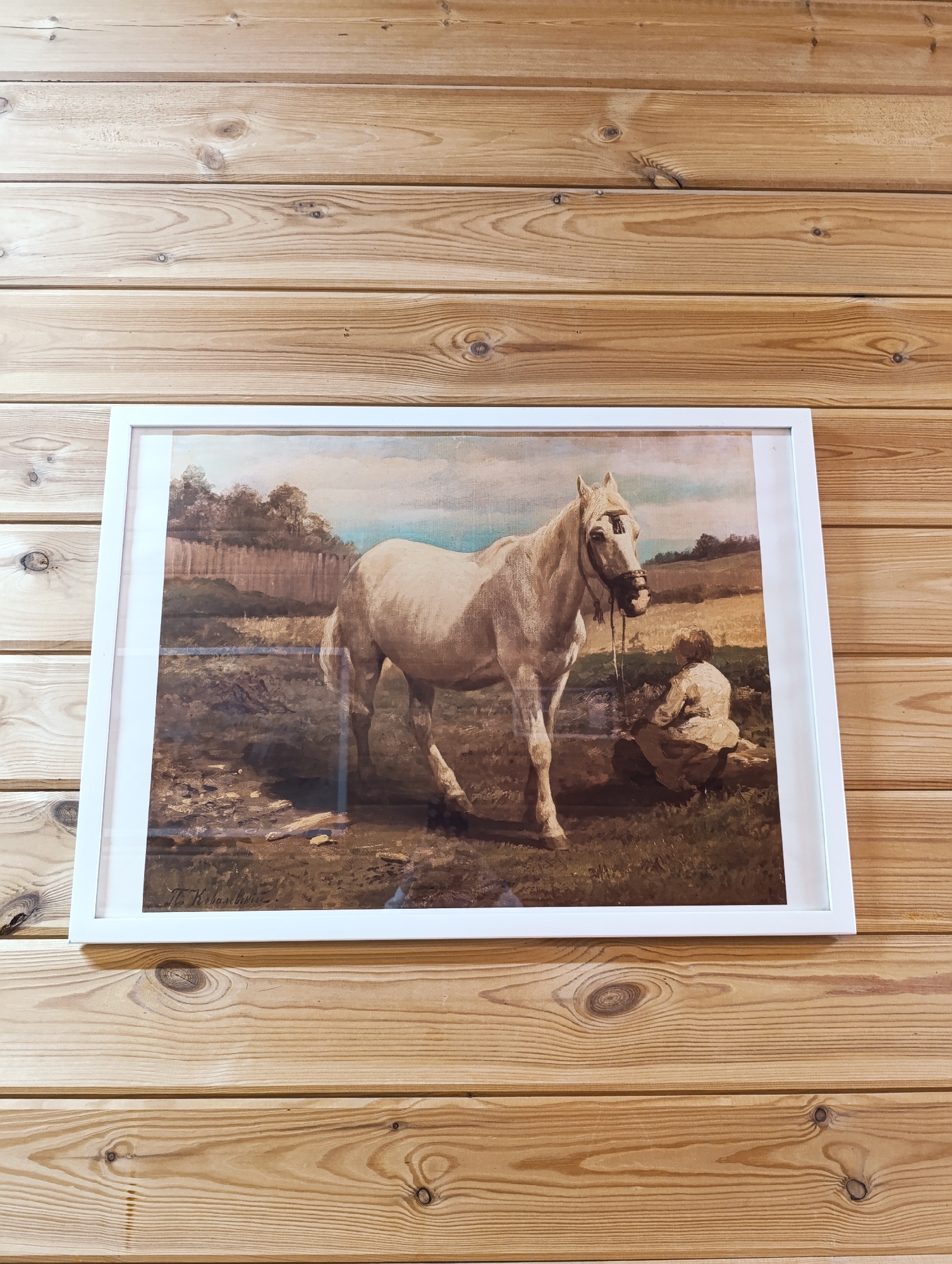 A framed painting of a white horse is hung on a wooden wall made of horizontal planks. The painting depicts the horse standing in a field with a cloudy sky and trees in the background. In the lower right corner of the painting, a person is sitting on the ground. The painting is signed in the lower left corner with the name "T. K. Baschitz." The frame is white and simple. The wooden wall has a natural wood grain and color.