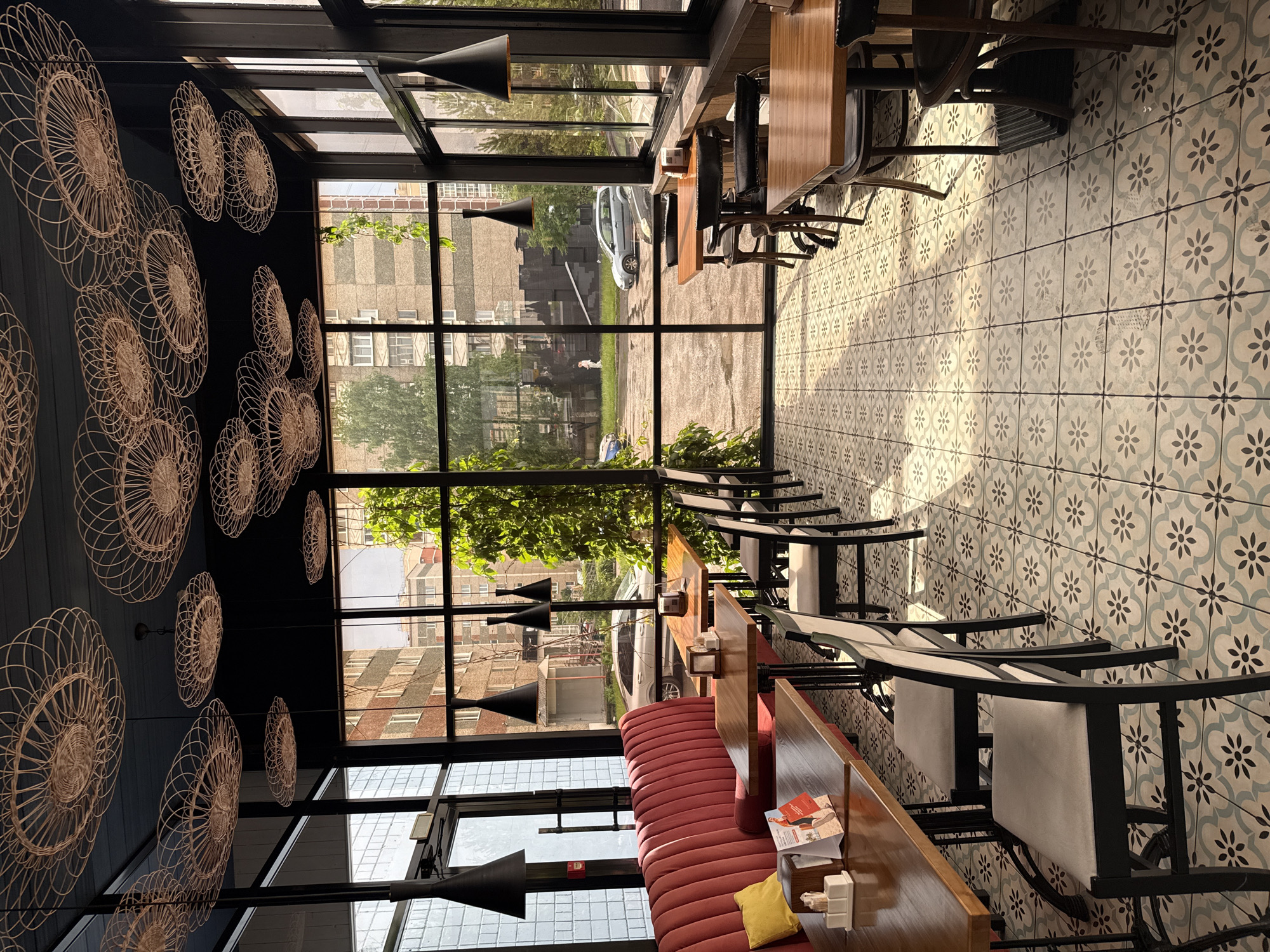 The image depicts the interior of a restaurant with large windows overlooking a city street in Izhevsk, Russia. The restaurant features a modern design with wooden tables and chairs, a red velvet booth, and decorative woven light fixtures suspended from the ceiling. The floor is covered in a patterned tile. Through the windows, buildings and cars are visible on the street, and some greenery suggests a residential area. The lighting indicates it is daytime, with sunlight streaming through the windows and casting shadows on the floor. There are no people clearly visible inside the restaurant, though a figure can be seen walking on the street outside.