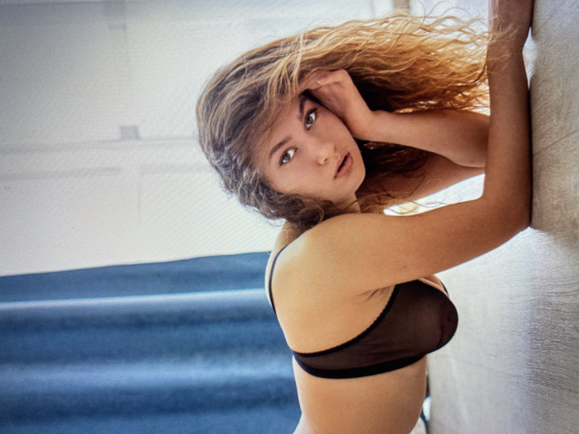 A woman with long, wavy blonde hair is captured in a provocative pose, leaning on her right arm. She is wearing a sheer black bra. Her head is tilted back, and her left hand is touching her hair near her forehead. Her eyes are open and looking towards the camera with a curious expression. The background is divided into two distinct areas: on the left, there are blue drapes, possibly curtains, with soft folds. On the right, a textured, light-colored surface, likely a wall or fabric, is visible. The lighting suggests it could be daytime, with a soft, diffused light illuminating the scene. The overall mood is intimate and alluring. There is no visible text in the image.