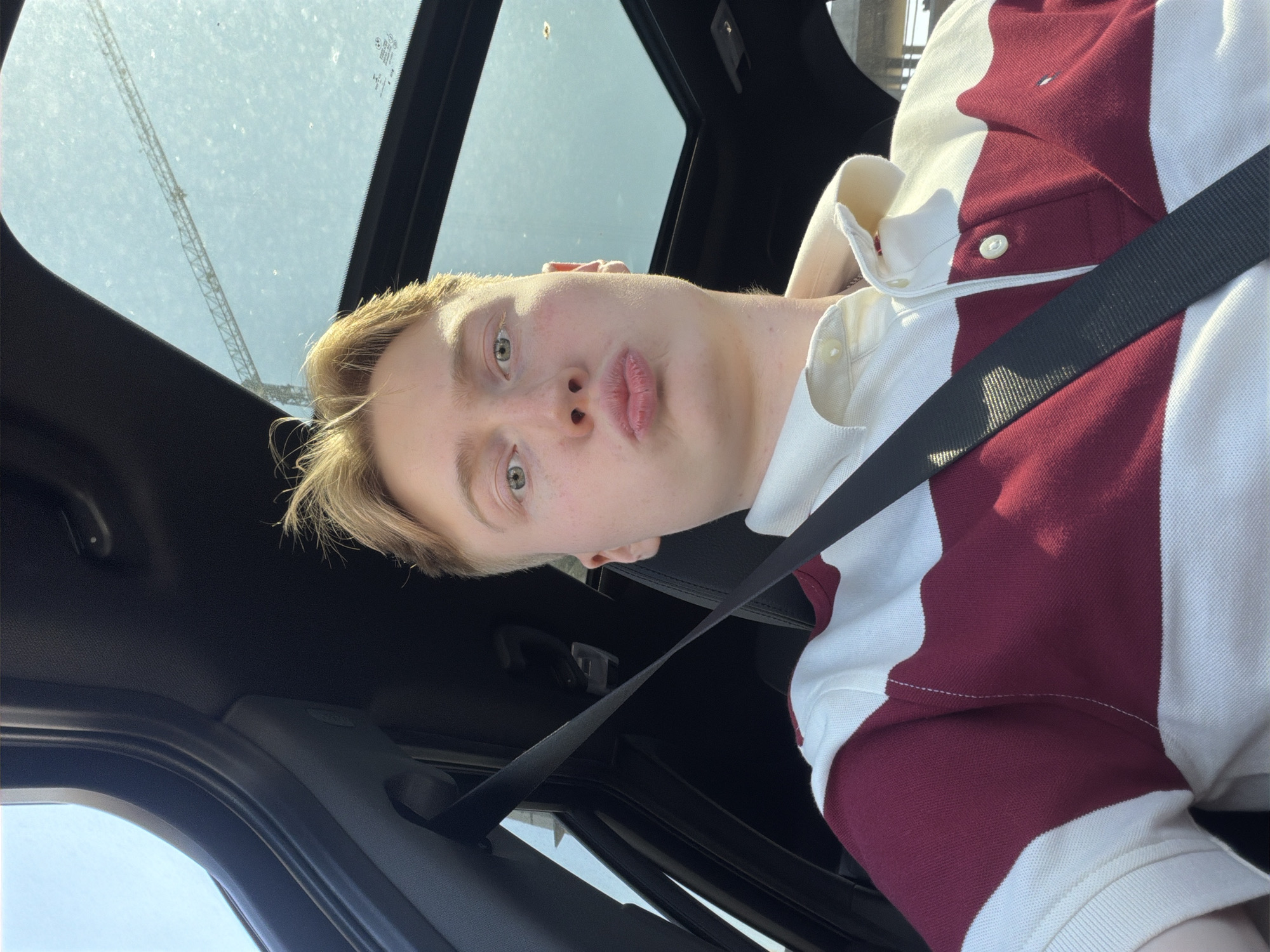 A young Caucasian male with blonde hair is seated in the passenger seat of a car, looking at the camera. He is wearing a white and maroon striped polo shirt and a black seatbelt is visible across his chest. The car has a sunroof, through which a crane and a bright sky are visible. The interior of the car is dark, with black upholstery and trim. The image appears to have been taken during the day, with natural light illuminating the scene. The specific location in the city of Kyiv, Ukraine, is not identifiable from the image.