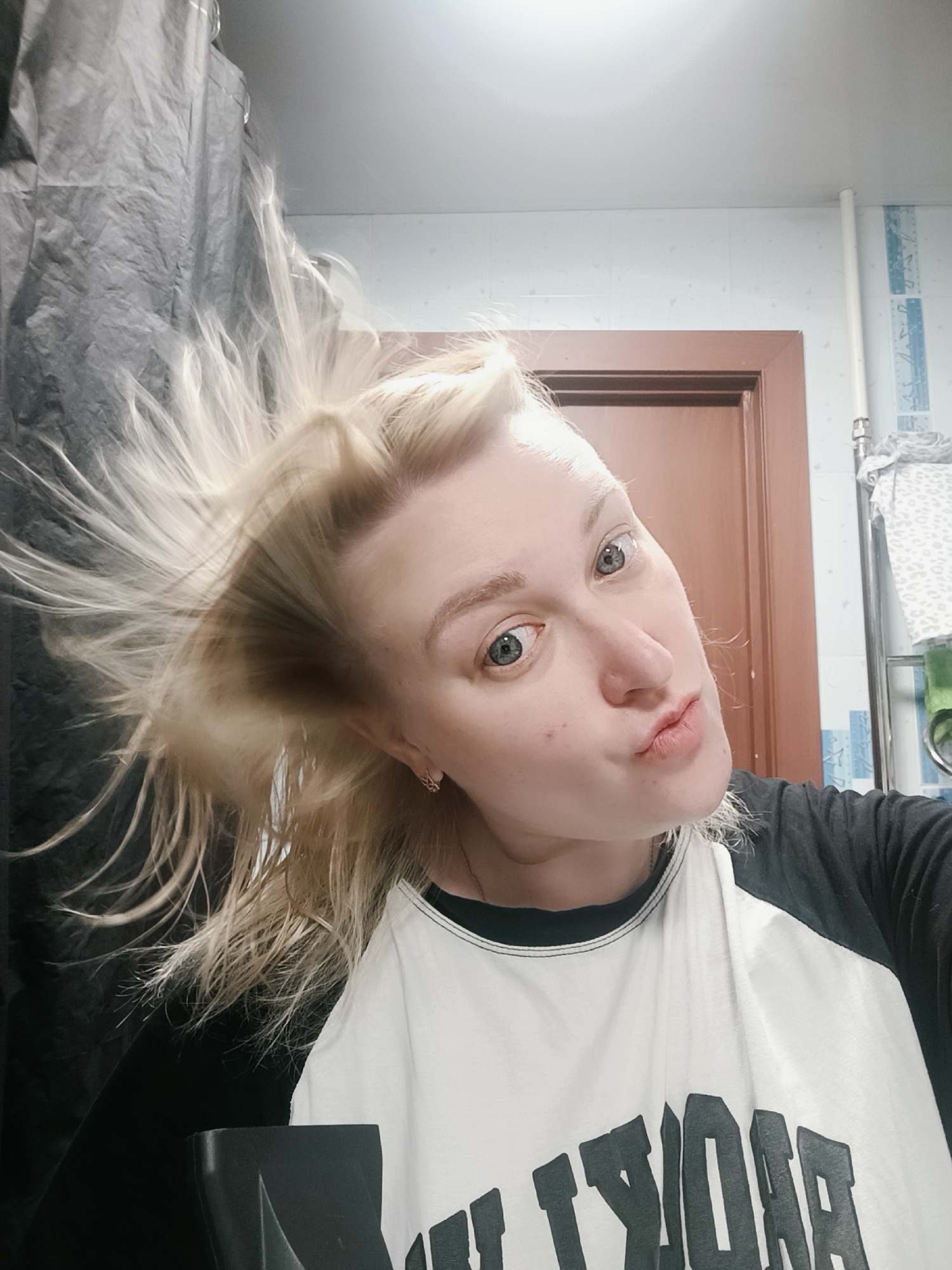 A woman with blonde hair is indoors, likely in a bathroom. Her hair is tossed to her left, partially obscuring a dark-colored shower curtain. She is wearing a black and white baseball-style shirt with large lettering. Behind her is a brown door and white tiled wall with a blue decorative pattern. A white pipe runs vertically along the wall. The woman is looking at the camera with pursed lips. The location is possibly in the city of Chaikovsky, Russia.