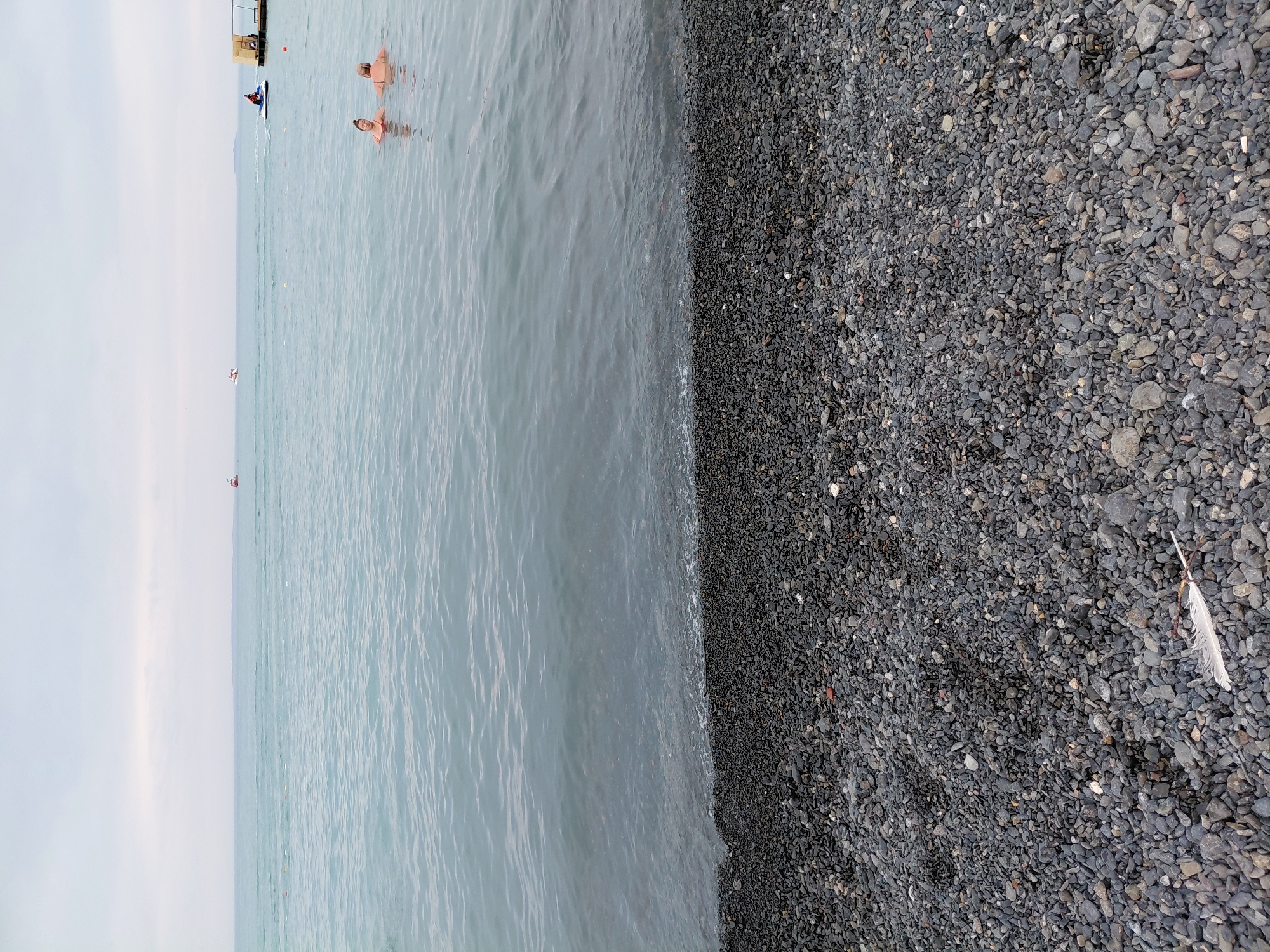 A wide shot shows a pebble beach in Ақши, Kazakhstan, with the calm, gray water of a large body of water in the background. Two people are partially submerged in the water, with their upper bodies visible. In the distance, a jet ski is being ridden by a person. Further out, two small boats and a larger vessel are present. A wooden pier extends from the right side of the frame into the water. The sky is overcast. A single white feather lies on the pebble beach in the foreground.