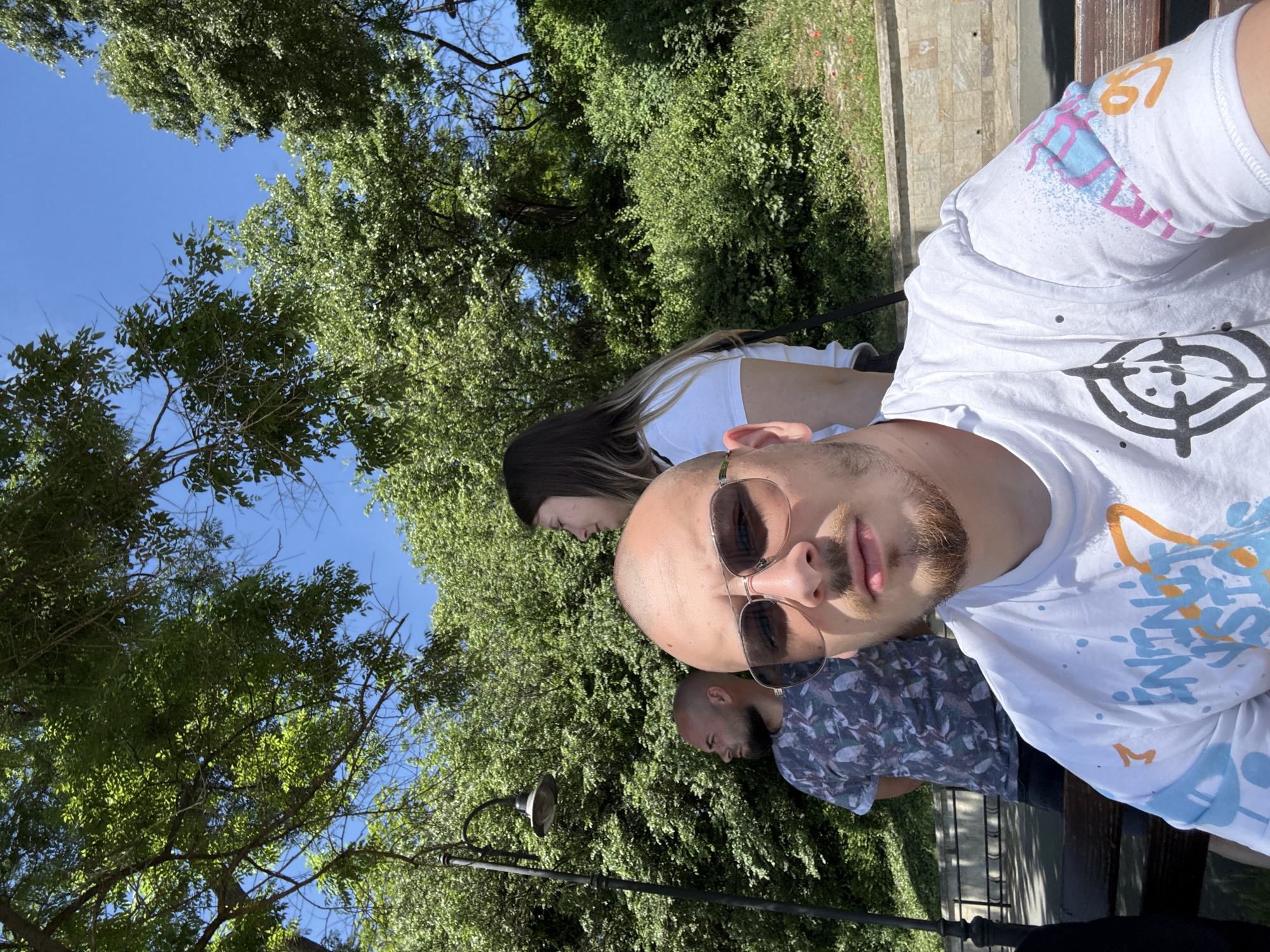 The image depicts three people, a bald man wearing sunglasses taking a selfie. Behind him to the left is a woman with long brown hair and to the right another man wearing a patterned shirt. They appear to be sitting on a bench in a park setting, with lush greenery and trees visible in the background and a clear blue sky. Based on the visual characteristics, this image was likely taken in Burgas, Bulgaria.